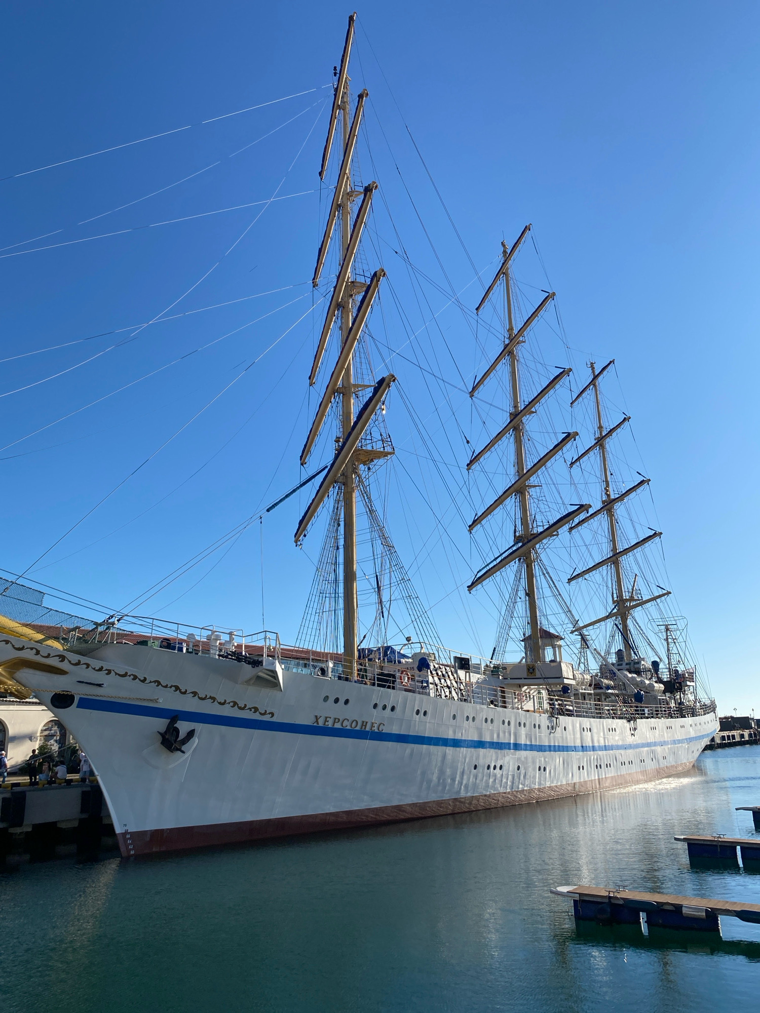 A large white sailing ship, identified by the Cyrillic lettering "ХЕРСОНЕС" on its hull, is docked in a harbor under a clear blue sky. The ship has four masts with numerous rigging lines. Several people are visible on the dock to the left of the ship, and other smaller floating structures are in the water. The ship's hull displays draft markings. This scene is located in the city of Sochi, Russia.