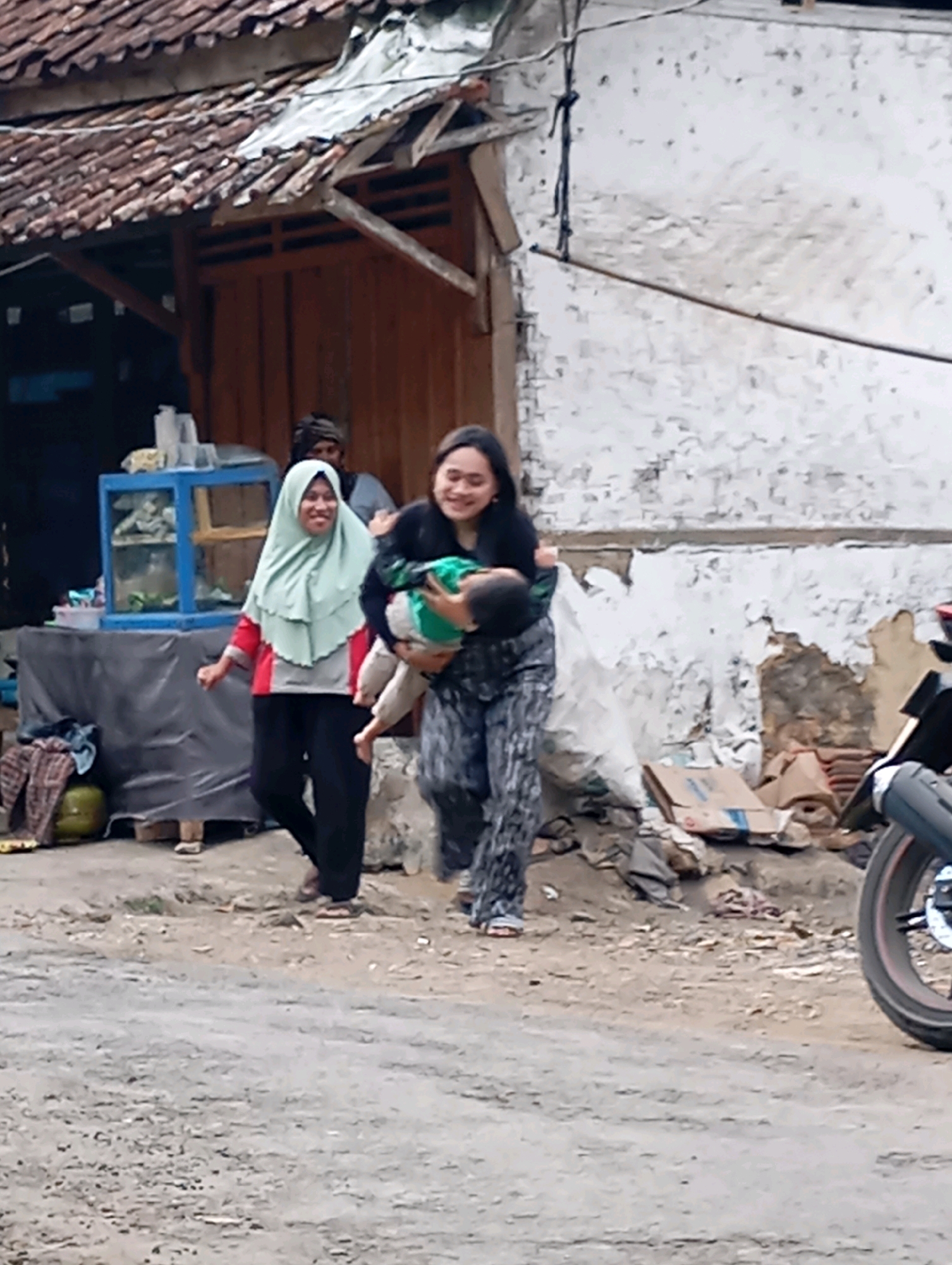 In the city of Cigunung, Indonesia, two women are walking on a dirt road. The woman on the left wears a light green hijab, a red and gray shirt, and black pants. She is smiling and looking towards the right. The woman on the right, holding a baby, wears a black shirt and patterned pants. She is also smiling and looking towards the baby she is carrying. The baby is wearing a green outfit. Behind them, a building with a tiled roof and wooden siding is visible. To the left of the building, a blue glass display case sits on a table covered with a gray tarp. Various items, including a green gas tank and a folded blanket, are near the table. The environment appears to be a street or alley with debris, including cardboard boxes and plastic bags, scattered around. A motorcycle is parked on the right side of the image.