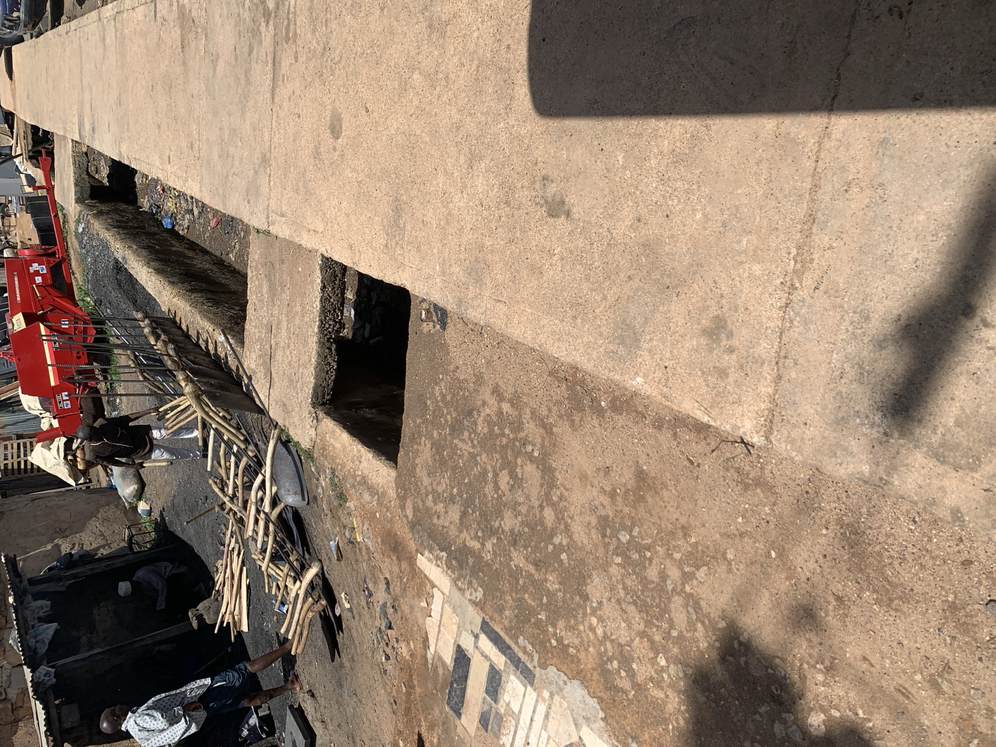 This image captures an active outdoor workshop or market scene in Gura Top, Nigeria, viewed from a slightly elevated vantage point. The time of day appears to be midday or early afternoon, judging by the strong, distinct shadows indicating bright sunlight.
Several people are visible, engaged in various tasks. In the foreground, a man wearing a light-colored, possibly patterned shirt and dark shorts is positioned near the bottom edge of the frame. Further into the scene, another man in a dark shirt and white trousers stands by a prominent red machine. This machine, possibly agricultural equipment, clearly displays the text "BAMFORD R" on its side. Near him, piles of wooden sticks or poles are neatly arranged, suggesting they are raw materials for work.
Below the main ground level, in a recessed and darker area, at least two more individuals are observed working. One figure is distinguishable by a white cap, indicating a workshop or activity taking place in a pit or lower structure. The ground in this section is dark and appears stained, possibly by charcoal or heavy usage.
The scene is bordered on the right by a substantial concrete structure, likely a walkway or part of a bridge. This structure features several open, rectangular cutouts, which may function as drainage channels or access points. The ground is unpaved, consisting of dirt and scattered debris, including discarded plastic. On the lower right portion of the concrete structure, broken ceramic tiles displaying a black, white, and grey geometric pattern are partially embedded in the surface. The overall setting conveys an industrious and bustling environment.