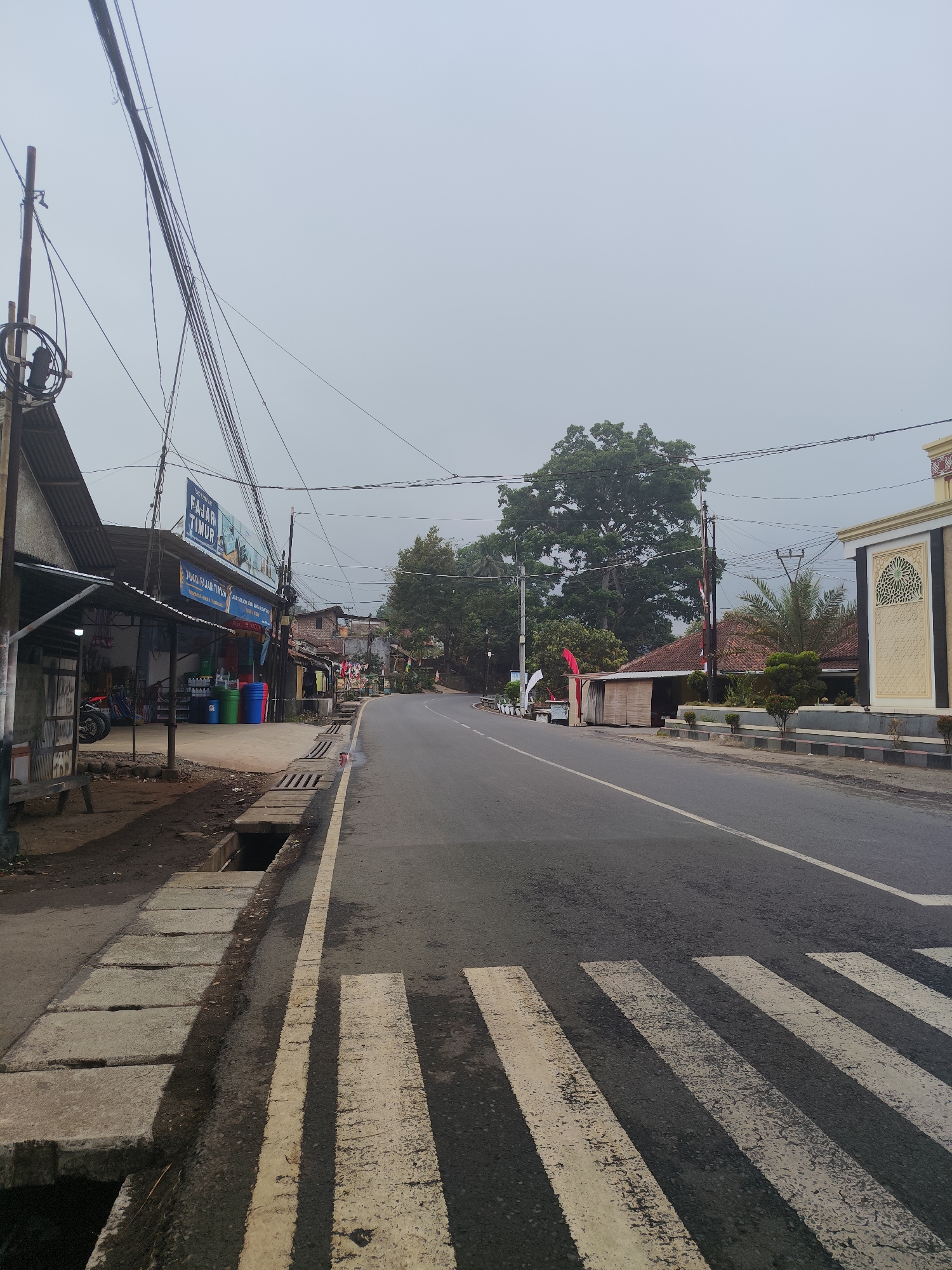 The image depicts a roadside view in Tasikmalaya, Indonesia, on a hazy or overcast day. The foreground features a pedestrian crossing with white stripes painted on the asphalt road. To the left of the road, a sidewalk is visible with concrete slabs and a drainage channel. Adjacent to the sidewalk is a small building with a corrugated metal roof, identified by a blue sign that reads "FAJAR TIMUR". This establishment appears to be a store, with various goods like plastic containers displayed outside. Further down the road, to the right, is a building with ornate yellow and white detailing, possibly a religious institution or public building. The road curves to the left and is lined with trees and smaller structures. Power lines are strung across the scene, supported by poles. Several Indonesian flags are also visible, attached to poles and buildings. No people are clearly discernible in the image. The overall impression is of a typical street scene in a town or rural area in Indonesia.