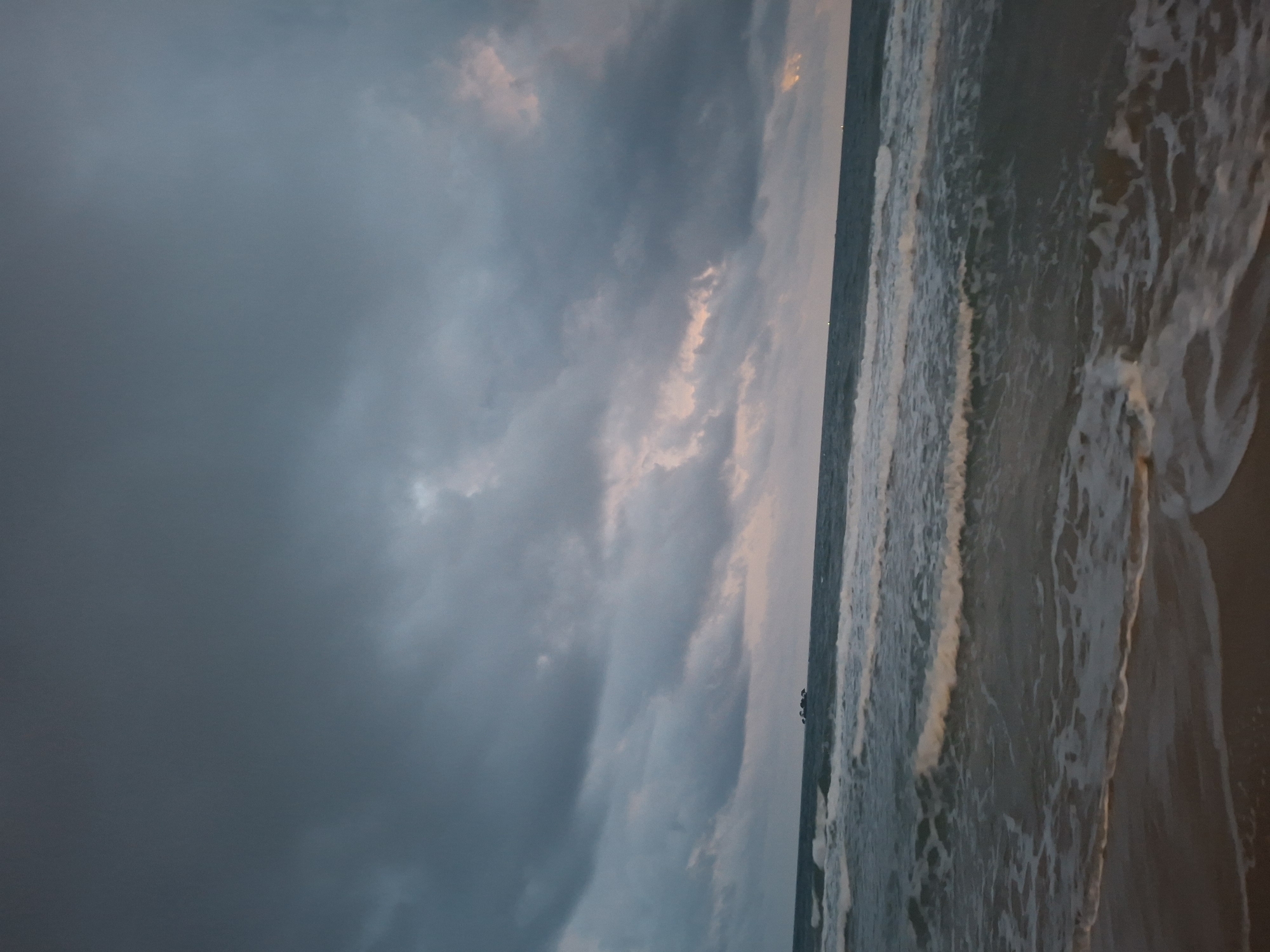 The image depicts a wide shot of a beach at dusk, with heavy, dark clouds dominating the sky. The ocean is visible in the lower half of the frame, with gentle waves rolling onto the sandy shore. There are no people or visible structures in the frame, except for a faint, distant cluster of dark shapes on the horizon, possibly rocks or small islands. The overall mood of the image is somber and tranquil, with the muted colors and the approaching darkness contributing to a sense of quietude. The scene appears to be set on a tropical beach, likely in Phú Quốc, Vietnam, given the overcast sky and the tropical feel of the vegetation implied by the distant landmasses.