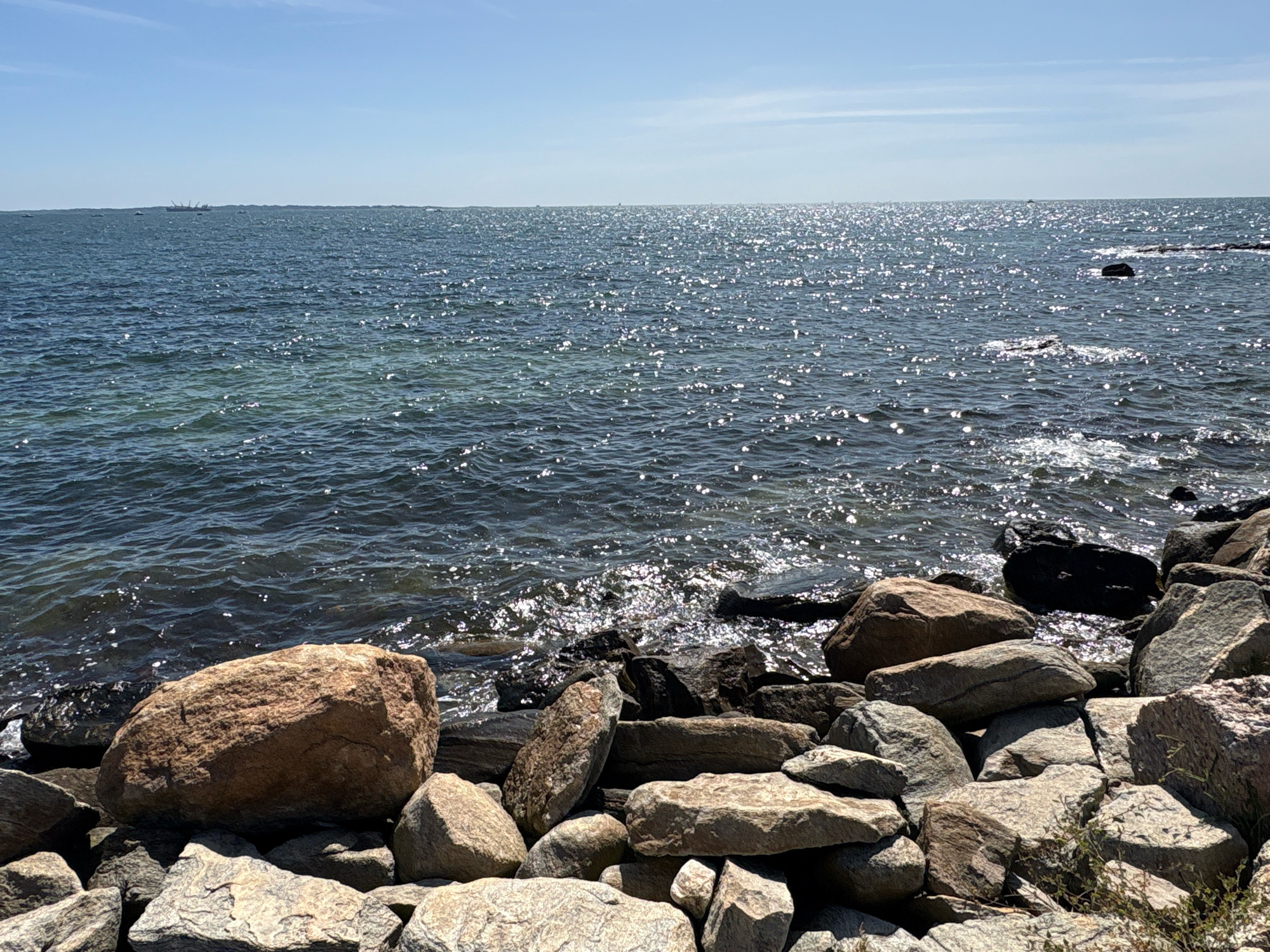 The image shows a rocky shoreline on a clear, sunny day. The ocean stretches to the horizon, with sunlight sparkling on the water's surface. In the distance, a large ship is visible on the water, along with several smaller boats. The foreground is dominated by a collection of large rocks and stones of various shapes and sizes, some of which are covered in moss or small plants. The sky is a bright blue with a few wispy clouds. The location is the coast of Waterford, United States of America.