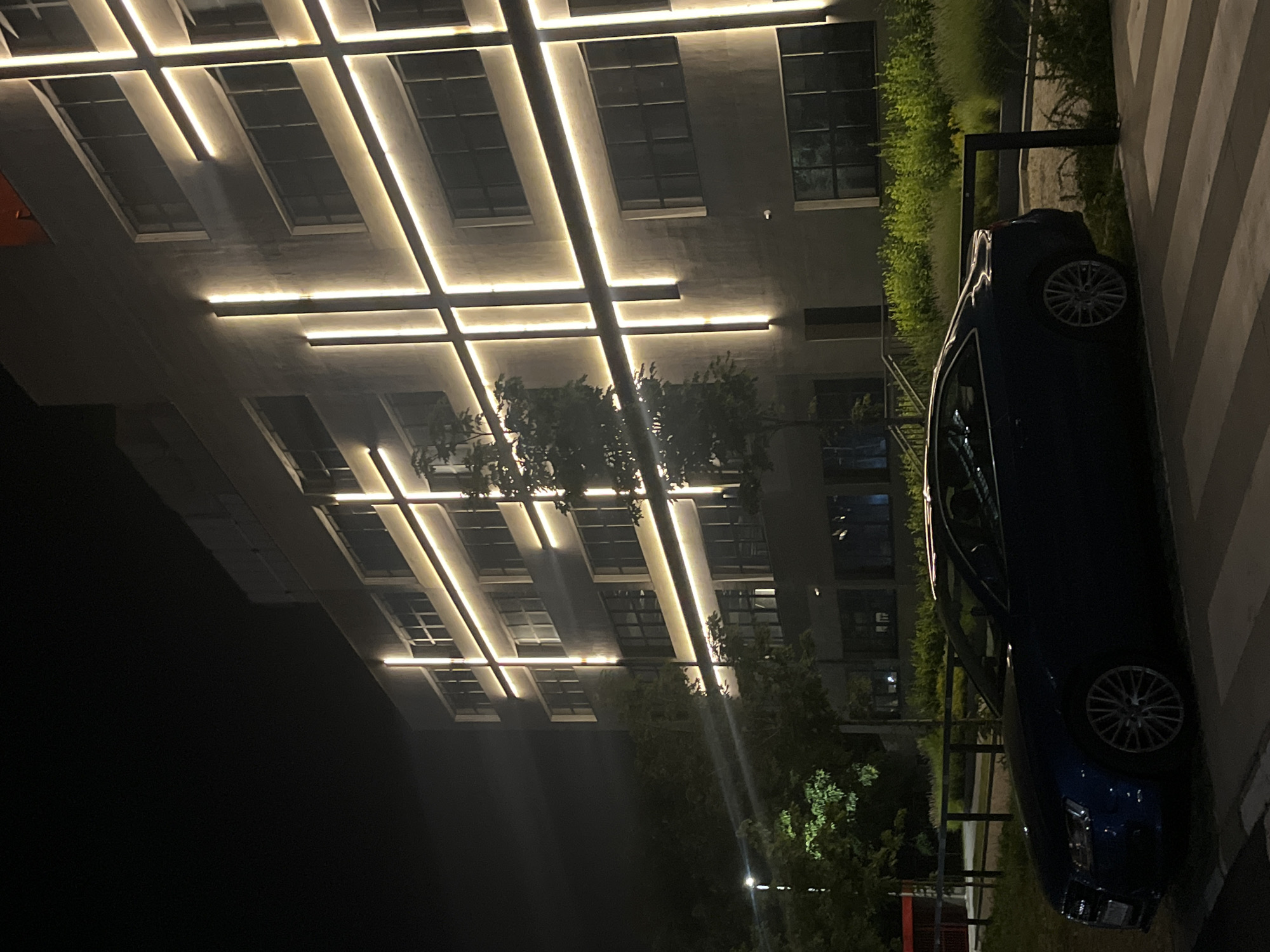 The image shows a dark blue coupe parked on a striped pavement in front of a modern building at night. The building's facade is illuminated by vertical and horizontal strips of warm-toned light, creating a grid pattern. There are several windows visible on the building. Lush green vegetation and trees surround the building and parking area. A black metal railing runs along the edge of the vegetation. The sky is dark. The scene appears to be an exterior shot in a city. Based on the context of other images associated with this scene, the location is likely Belgrade, Serbia, specifically the building known as the "Big Apple" (Veliki Zgrada) in New Belgrade, located at Bulevar Mihaila Petrovića 35.