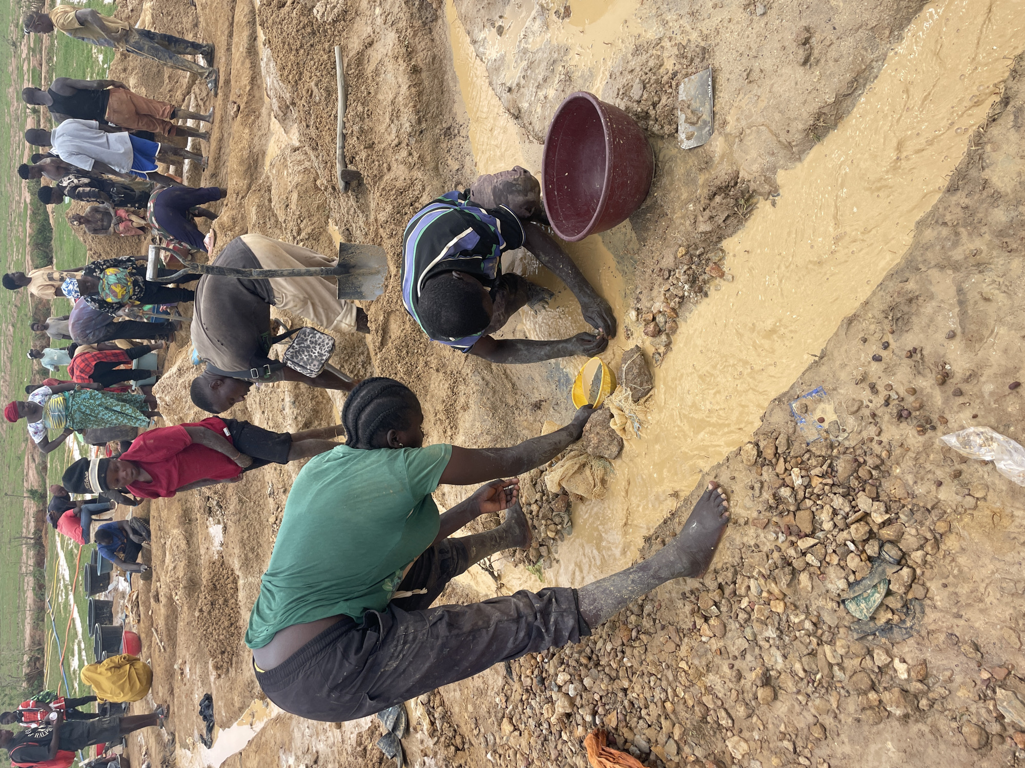 The image shows a group of people engaged in artisanal mining activities in Maiyanga, Nigeria. The scene is a muddy, rocky area with a shallow stream of dirty water flowing through it. Several individuals are working along the water's edge, some digging, others sifting through the sediment, and one using a yellow scoop. There is a large, dark-colored bucket near one of the miners. Most people are barefoot or have mud-covered feet and legs. The individuals are wearing casual clothing. In the background, there is a larger group of people also involved in the mining operation, and some greenery or vegetation is visible in the far distance. There are several dark-colored buckets in the background as well. A shovel is visible nearby.