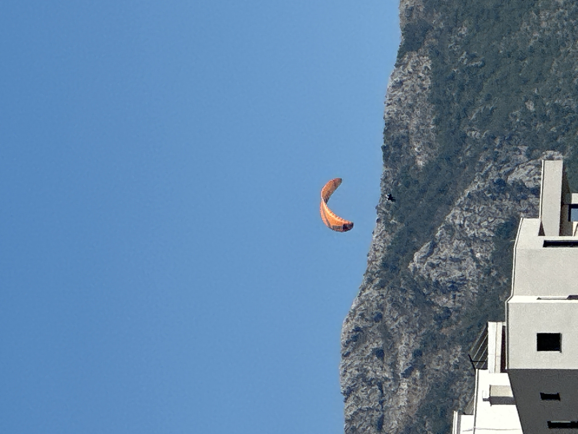 The image depicts a clear blue sky above a rugged, rocky mountain range. A bright orange paraglider is suspended in the air, with a small figure visible beneath it, presumably the pilot. In the foreground, a modern white building complex is partially visible. The scene is likely located near Boreti, Montenegro, given the characteristic mountainous terrain and coastal architecture.