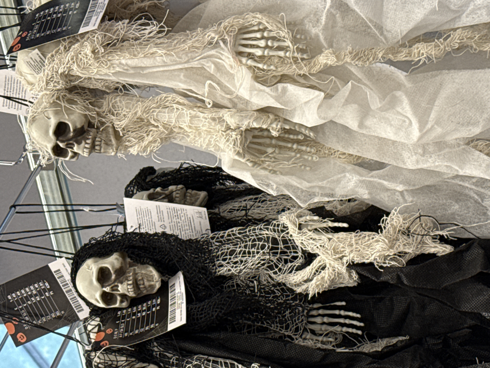 The image shows two Halloween decorations hanging on display. The decoration on the left is a skeleton with a black shroud and netting. The decoration on the right is a skeleton with a white shroud and netting. Both skeletons are hanging from a metal rack. The background is blurry and appears to be a store interior. The scene is indoors, likely a retail environment in Bad Lauterberg im Harz, Germany, given the price tags indicating different countries and currencies. The focus is on the Halloween decorations, with price tags also visible.