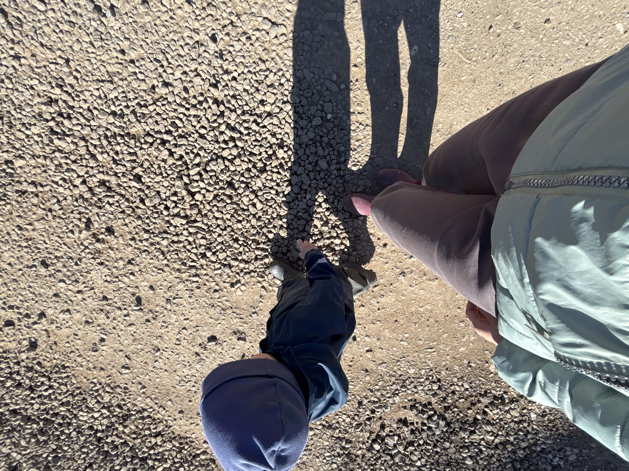 The image shows a top-down view of a person and a toddler standing on a gravel surface. The person is wearing brown pants, pink shoes, and a light green jacket. The toddler is wearing a blue hooded jacket and shoes. Shadows of two individuals are visible on the ground, suggesting a sunny day. The gravel surface is composed of small, irregularly shaped stones. The scene is likely outdoors in Ферма, Russia.