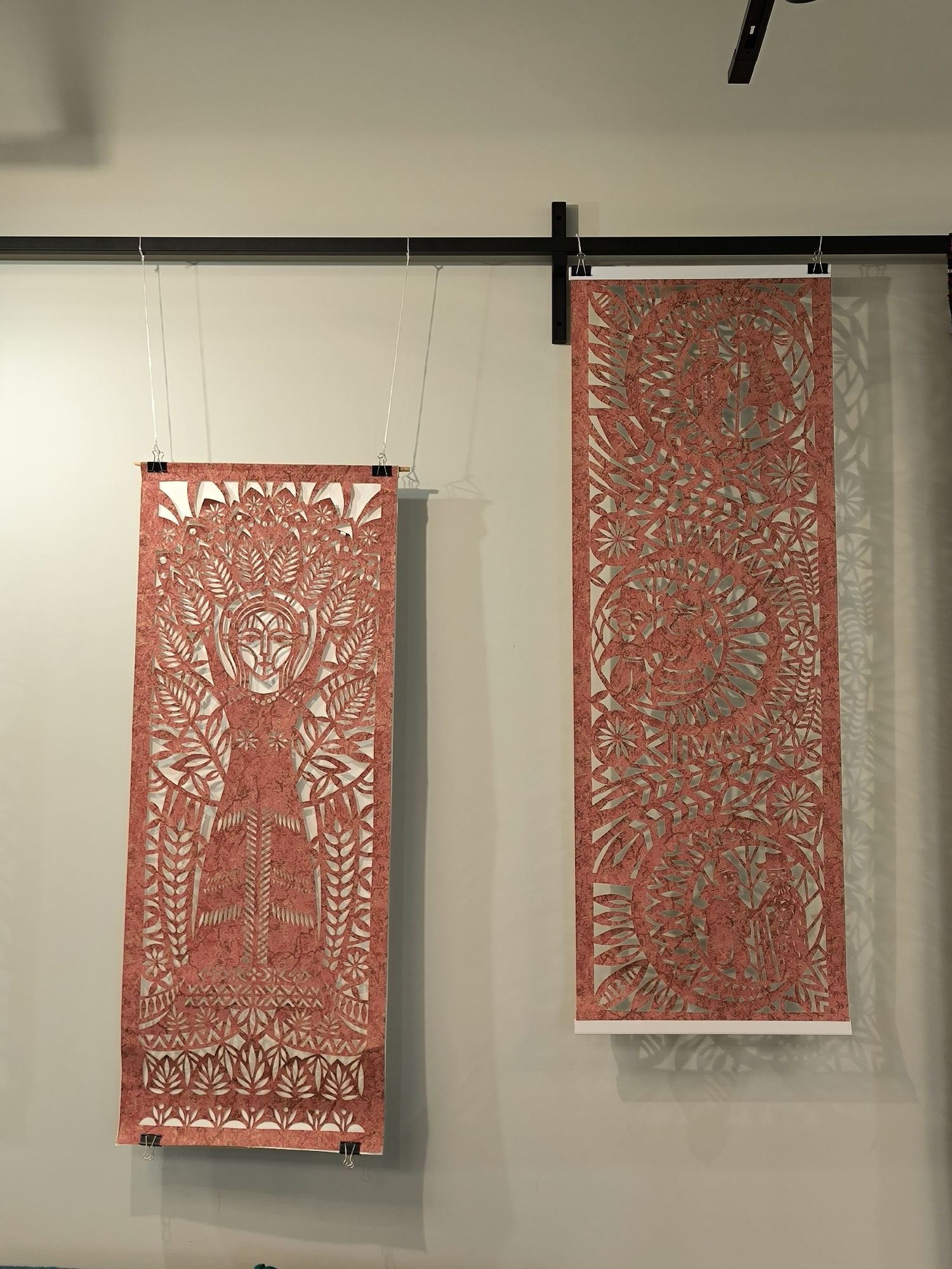 Two decorative art pieces hang on a wall in Vinnytsia, Ukraine. The pieces are rectangular, with intricate designs cut out of a textured reddish-brown material, possibly paper or thin corkboard, revealing the light-colored wall behind. The piece on the left depicts a central figure with foliage, possibly a woman with tree-like extensions around her head and body. The piece on the right features a complex, circular pattern with floral and geometric elements. Both pieces are suspended from a black metal track on the ceiling by thin, silvery wires attached to the top edges of the art with binder clips. The wall is a light, neutral color, and the lighting is bright and even.