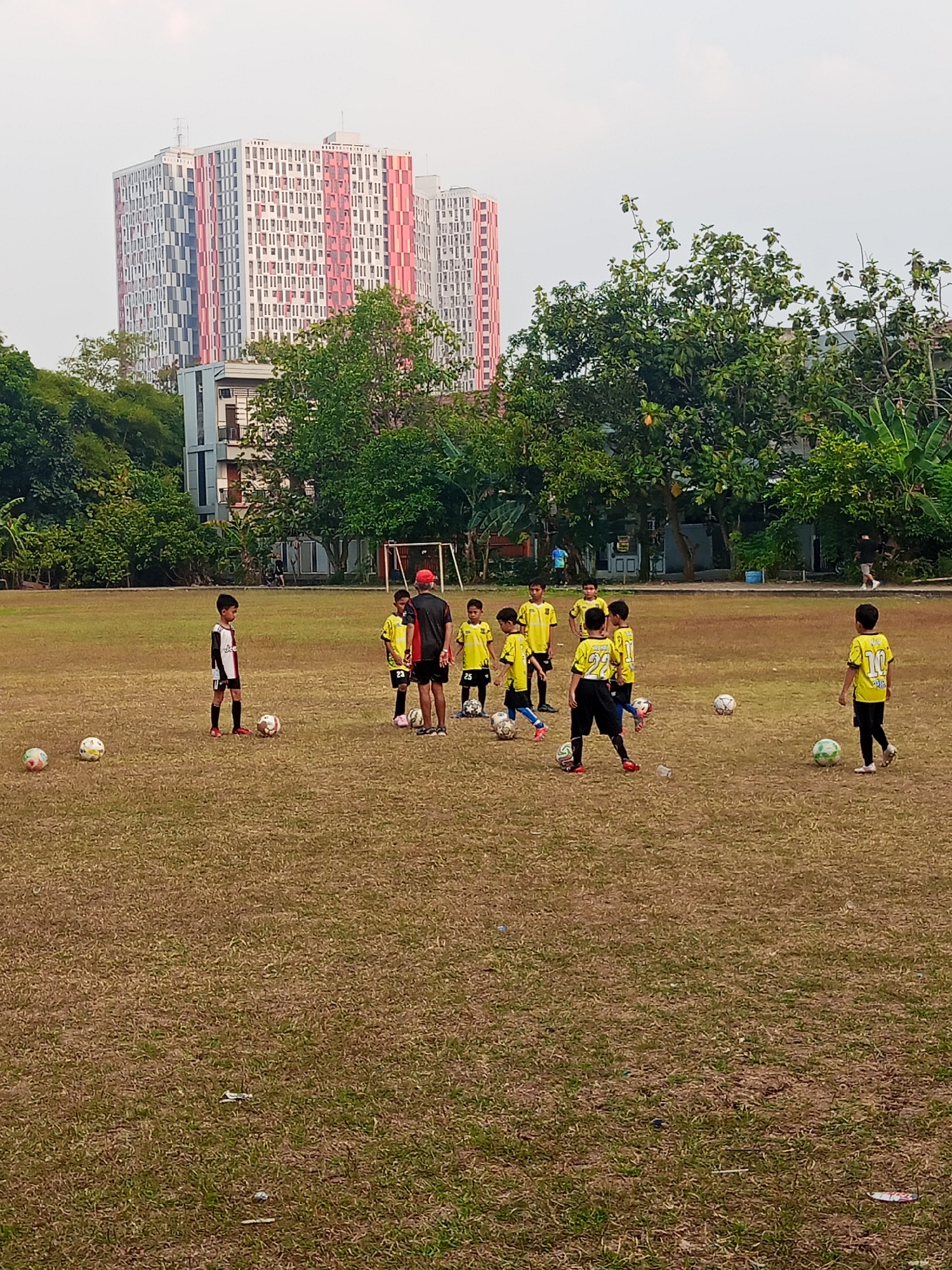 In Semarang, Indonesia, children in yellow and black soccer uniforms practice on a dry, brown grass field. There are several soccer balls scattered around the field. In the background, a tall, modern building with red and white geometric patterns stands against a pale sky. Lush green trees surround the field and the building. A soccer goal is visible in the distance.