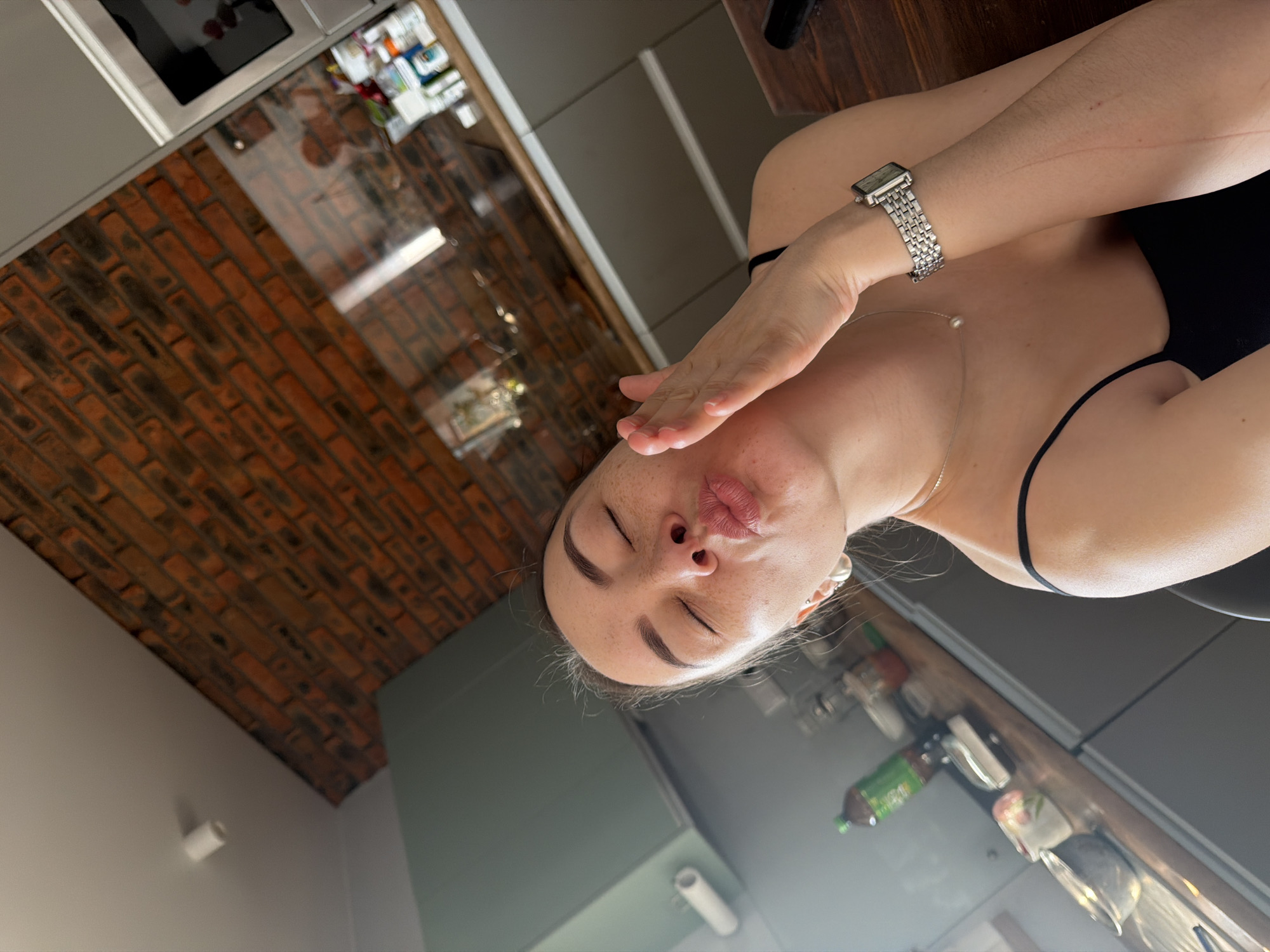 A selfie taken indoors shows a woman with closed eyes, a silver watch, and a silver necklace with a pearl. Her lips are puckered in a kissing gesture, and her right hand is raised near her face. She is wearing a black top with thin straps. The background consists of a modern kitchen with gray cabinets and a brick-patterned wall behind the cabinets. The scene likely takes place in an apartment or house. The photo does not have sufficient information to determine if it was taken in the city of Chaikovsky, Russia.