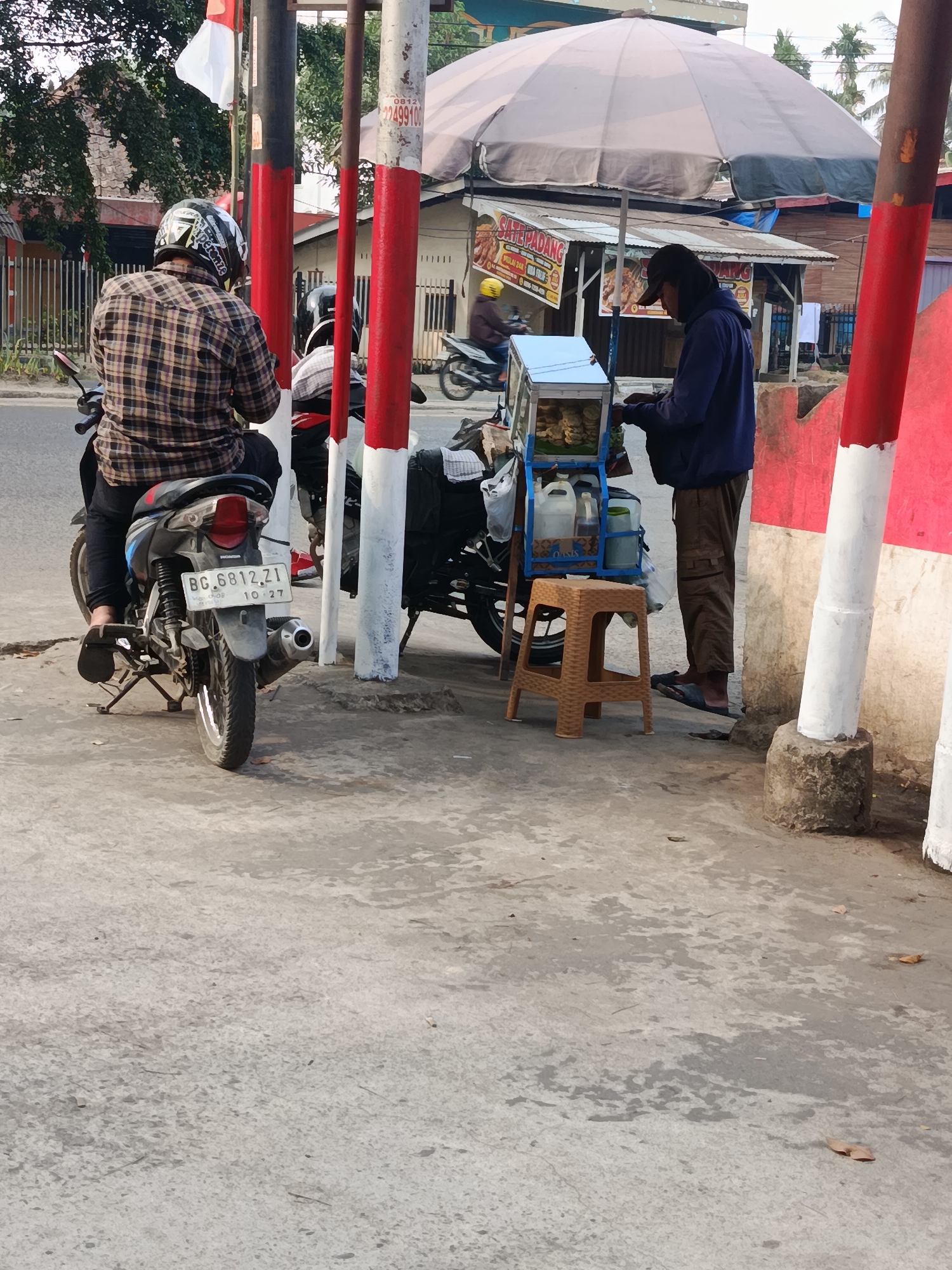 The image captures a street scene in Palembang, Indonesia. In the foreground, a person wearing a plaid shirt and a helmet sits on a motorcycle. To the right, another person in a dark hoodie and cap stands next to a food cart filled with food items and containers of liquid. A small plastic stool is placed in front of the cart. Behind the food cart, a large umbrella provides shade. In the background, buildings and trees are visible, with a glimpse of a red and white striped pole and a flag. The ground is paved with concrete. The license plate of the motorcycle reads "BG. 6812. ZI". The overall scene suggests a typical roadside food vendor operation.