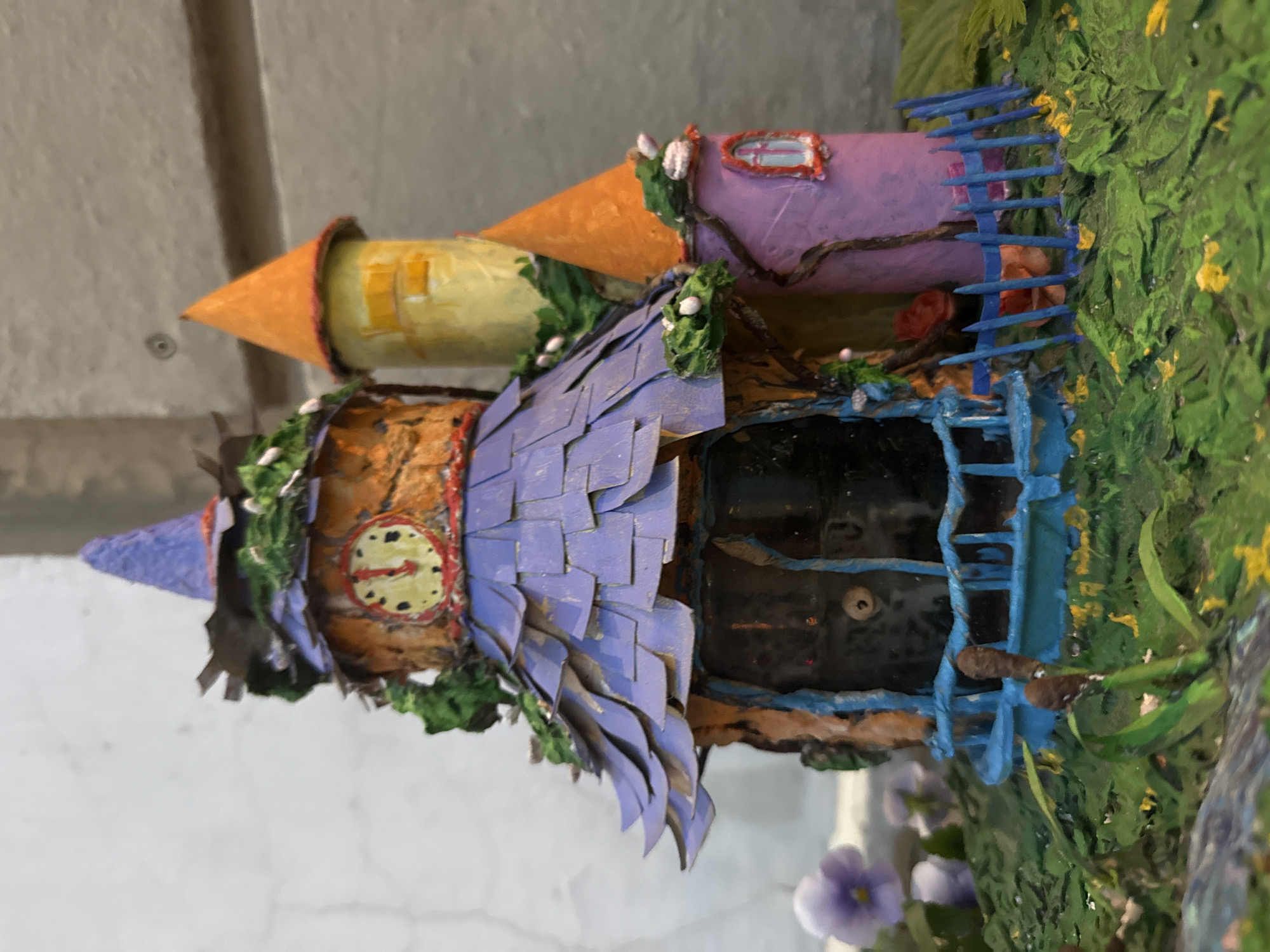 The image shows a whimsical, miniature fairy house or castle, crafted from various materials and painted in vibrant colors. The structure features multiple towers of different heights and colors, including a central purple-roofed section topped with a pointed blue hat. A clock face with red hands is prominently displayed on the orange-toned main body. Green foliage and small white flowers are artfully incorporated into the design, giving it a natural, overgrown appearance. A blue railing and a small blue picket fence surround the base of the structure. The ground around the fairy house is covered in textured green material, resembling grass and foliage, with scattered yellow accents and some purple flowers visible in the background. The backdrop appears to be a textured wall, possibly concrete or plaster, with subtle shadows suggesting it is outdoors. There are no people, animals, or specific identifiable landmarks of Saint Petersburg, Russia present in the image.