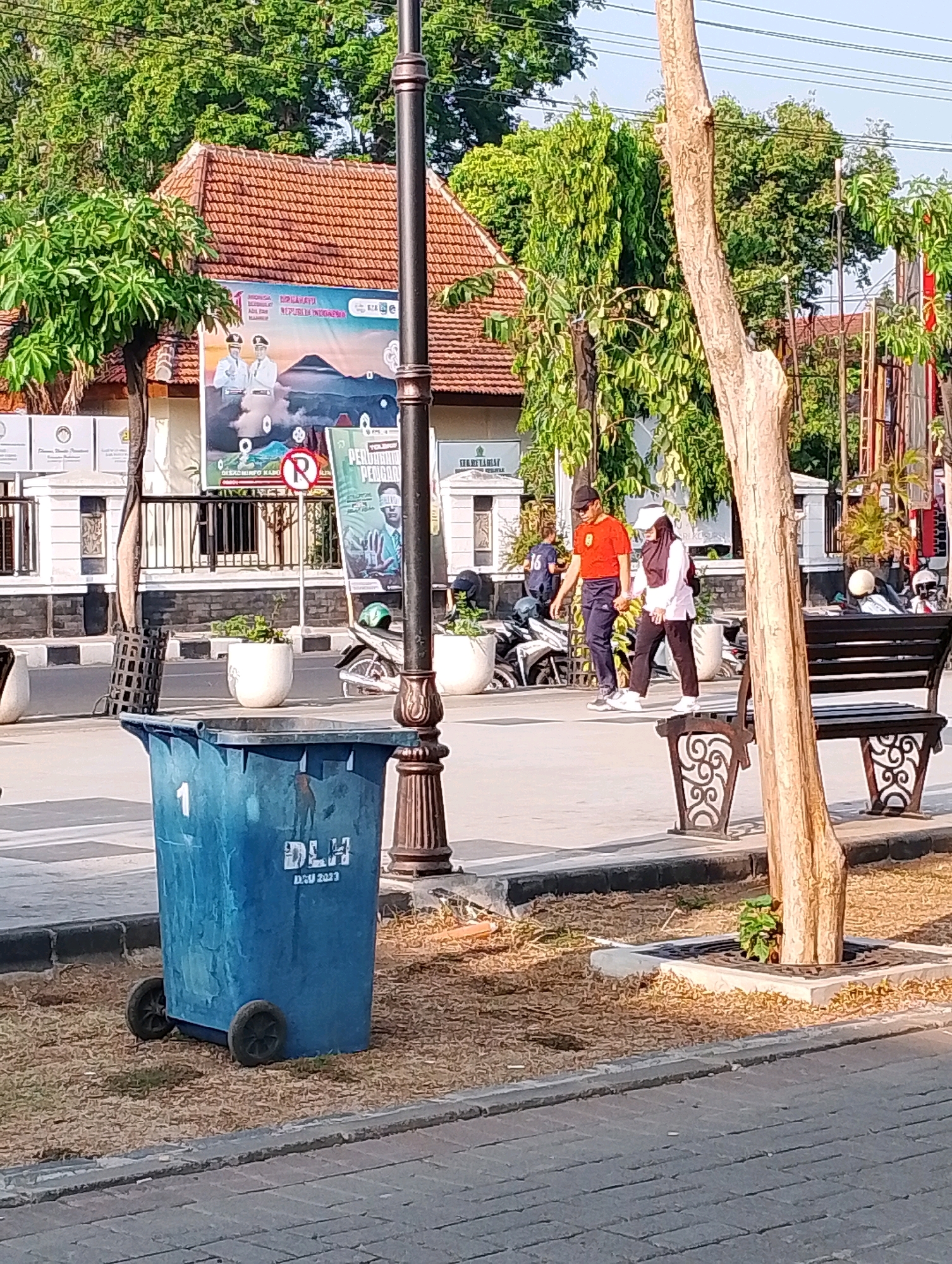 The image depicts a street scene in Probolinggo, Indonesia, likely a public area with a pedestrian walkway and adjacent buildings. In the foreground, a large blue trash bin with wheels is visible, marked with the number "1" and text that appears to be "DLH DGU 2023." To the right of the bin, a dark wooden park bench with decorative metal supports is placed near a young tree with sparse foliage.
Behind the bin and bench, a paved walkway stretches across the image. Two individuals are walking on this walkway. The person on the left is a male wearing a red t-shirt, dark pants, and a black cap. The person on the right is a female wearing a white long-sleeved top, black pants, and a white headscarf. They are walking side-by-side, in the same direction.
In the background, several elements suggest a bustling urban environment. A building with a red-tiled roof and white walls is prominent. A large banner with images of two men in suits and a mountain landscape is displayed on the building's facade. Other banners and signs are also visible. Several motorcycles are parked in the mid-ground, and trees with dense green foliage line the street, suggesting a well-vegetated urban area. Streetlights and utility poles are also present. The overall lighting indicates it is daytime, likely morning or afternoon, with sunlight casting shadows. The scene appears to be a public space designed for pedestrian use and sanitation.