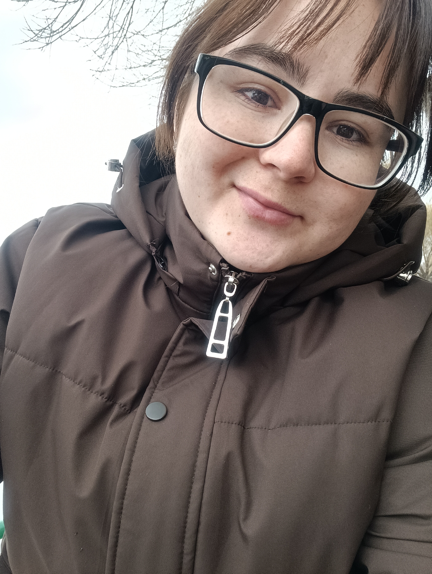 A selfie of a young woman wearing large, black-framed glasses and a brown puffer jacket with a hood. The jacket is partially zipped, revealing a metallic zipper pull with a rectangular cutout design. A single black button is visible on the jacket's left side. The woman has fair skin with freckles and bangs. The background is blurred but appears to show branches of bare trees against a pale sky. Based on context clues, it is implied she is located in the city of Chaikovsky, Russia.