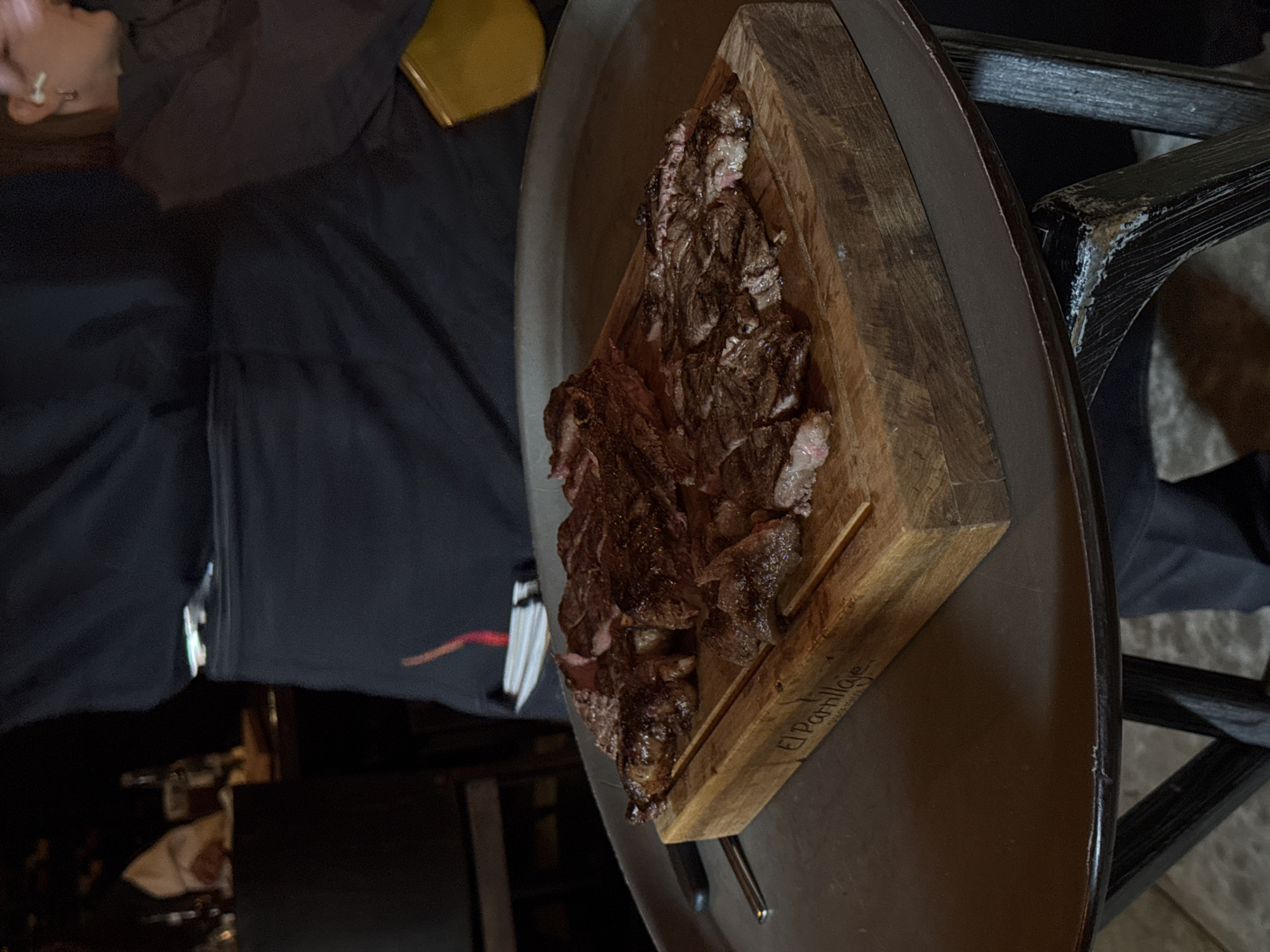 The image depicts a serving of grilled meat, likely beef, presented on a wooden board atop a dark, circular table. The meat is sliced and appears medium-rare, with visible grill marks. The wooden board has the inscription "El Parrilla" followed by smaller text underneath, suggesting it is the name of the restaurant. The table surface is dark brown or black and reflects light, indicating a glossy finish. In the background, there are glimpses of a person wearing dark clothing, and a dimly lit interior, likely a restaurant. The setting suggests the city of Puebla, Mexico, based on the restaurant name "El Parrilla." The atmosphere appears to be upscale dining.
