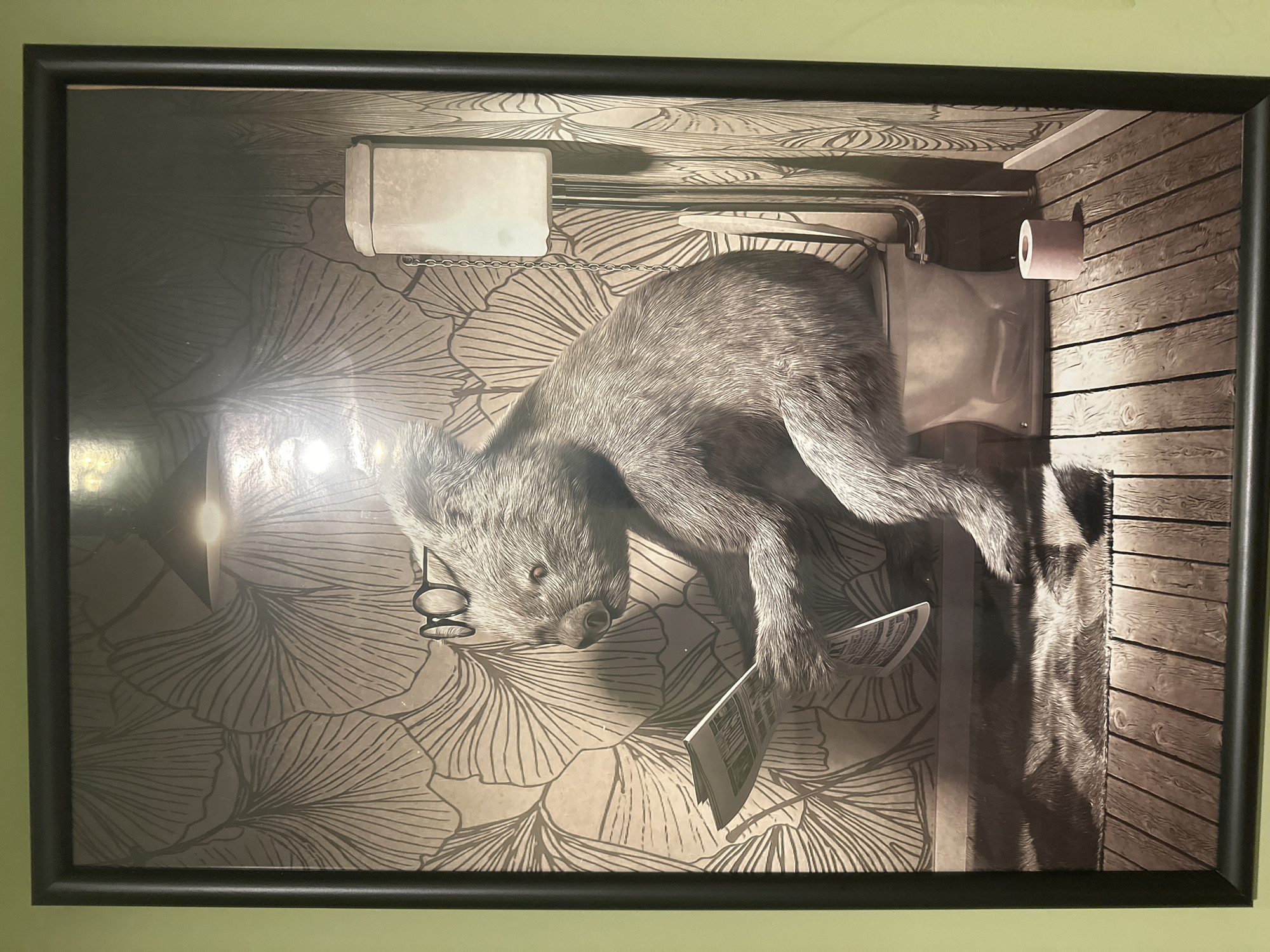 The image shows a framed print of a furry animal, possibly a koala, wearing glasses and reading a newspaper while sitting on a toilet. The toilet is located in a bathroom with floral wallpaper and wooden floors. The location in Sofia, Bulgaria is not visible in this image.