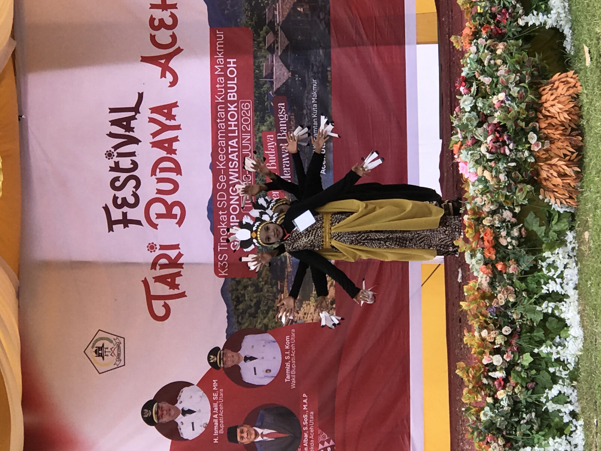 This image depicts an outdoor cultural performance likely in the city of Langkuta, Indonesia, as suggested by the text on the banner. In the foreground, a young female performer, dressed in traditional attire including a yellow sarong and a patterned bodice, is captured mid-dance with her arms extended and hands in a graceful pose. She is adorned with a distinctive headdress. Behind her, a large banner serves as the backdrop, featuring bold text that reads "FESTIVAL TARI BUDAYA ACEH" (Aceh Cultural Dance Festival) and further details about the event: "K3S Tingkat SD Se-Kecamatan Kuta Makmur Simpong Wisata Lhok Buloh 10-13 JUNI 2026". The banner also displays images of individuals in formal attire, identified as officials, with names and titles such as "H. Ismail A Jalil, SE, MM Bupati Aceh Utara" and "Tarmizi, S.I. Kom Wakil Bupati Aceh Utara". A logo is visible in the upper left corner of the banner. The stage is decorated with a dense arrangement of artificial flowers and foliage. The overall scene suggests a lively community event celebrating local culture and heritage.