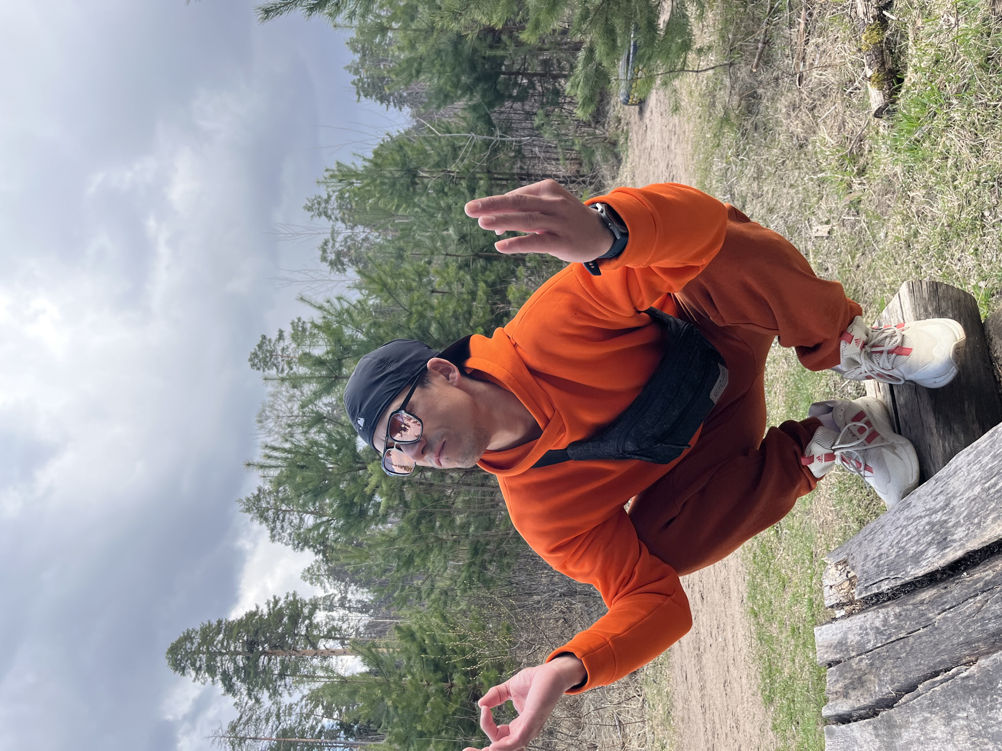 This outdoor portrait captures a man crouching or sitting on a weathered wooden plank, set against a backdrop of pine trees and an overcast sky. The image appears to be taken in a natural area, likely a forest or park, consistent with the location context of Chaikovsky, Russia.
The subject is a man wearing a bright orange hooded sweatshirt, matching orange sweatpants, white sneakers with red stripes (Adidas brand visible), dark sunglasses, and a black baseball cap with a white "NY" logo (New York Yankees). A black fanny pack or sling bag is worn across his chest. He also has a watch on his left wrist.
He is positioned with his body angled, looking towards the right side of the frame with a somewhat neutral expression. His left hand is raised, forming a "shaka" or "hang loose" gesture, while his right hand rests closer to his body, forming a mudra-like "ok" gesture with thumb and forefinger touching. This pose suggests a moment of contemplation, relaxation, or a stylized photo opportunity.
The background features numerous pine trees of varying heights, indicating a wooded environment. A dirt path or clearing is visible behind him. The sky is largely overcast, dominated by light grey clouds, suggesting a cloudy daytime. The ground around the wooden structure is covered with sparse grass and dirt. The overall impression is one of a casual outdoor setting.
