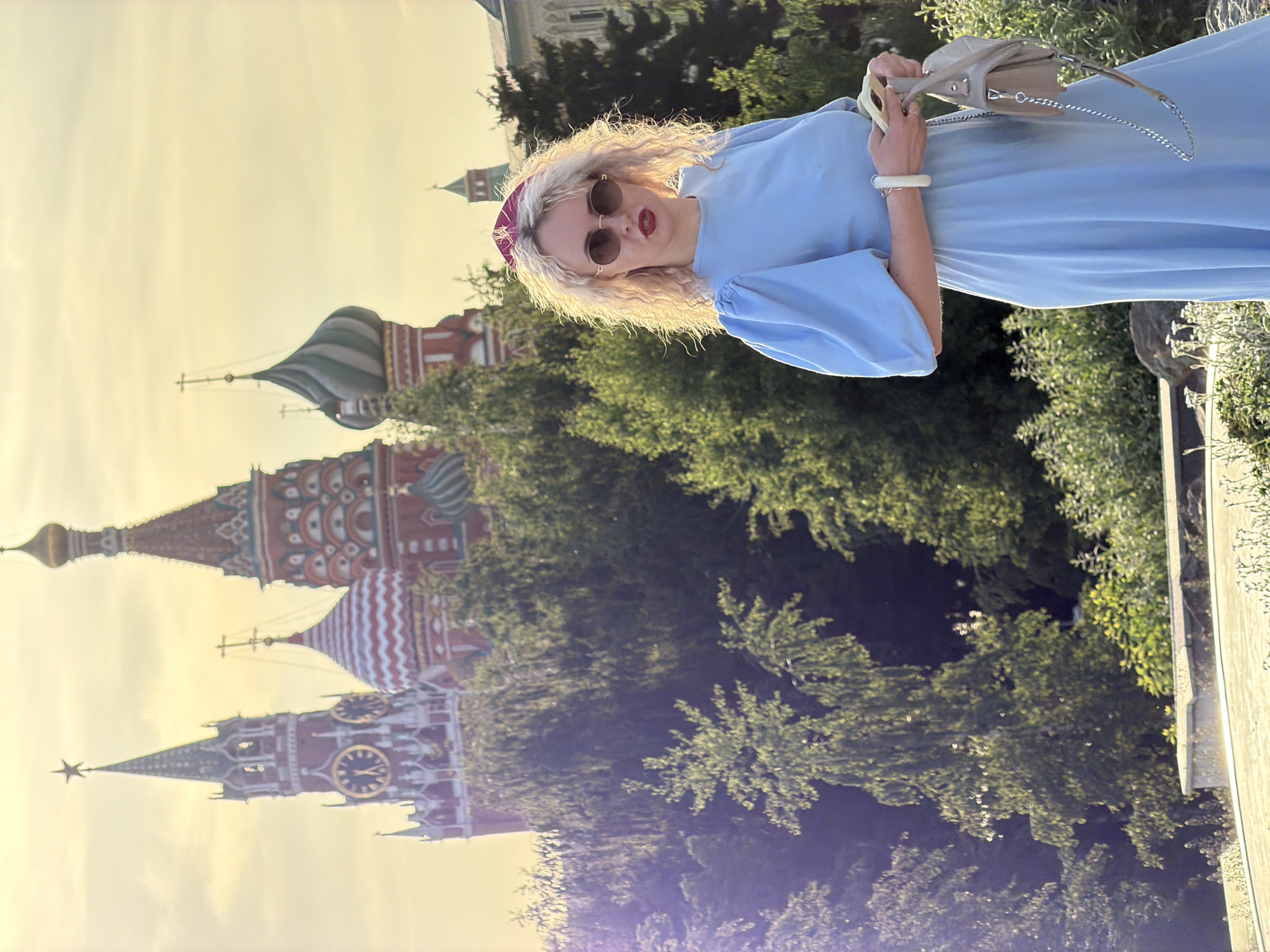 A medium shot of a woman standing near the Moscow Kremlin in Moscow, Russia. The woman, who has blonde curly hair and is wearing a light blue dress with puff sleeves, stands on the right side of the frame. She is holding a beige purse. The Kremlin buildings, including the towers of the Red Square, are visible behind a dense line of green trees. The sky is a soft, hazy yellow, indicating either sunrise or sunset. The orientation of the photograph is rotated 90 degrees counterclockwise.