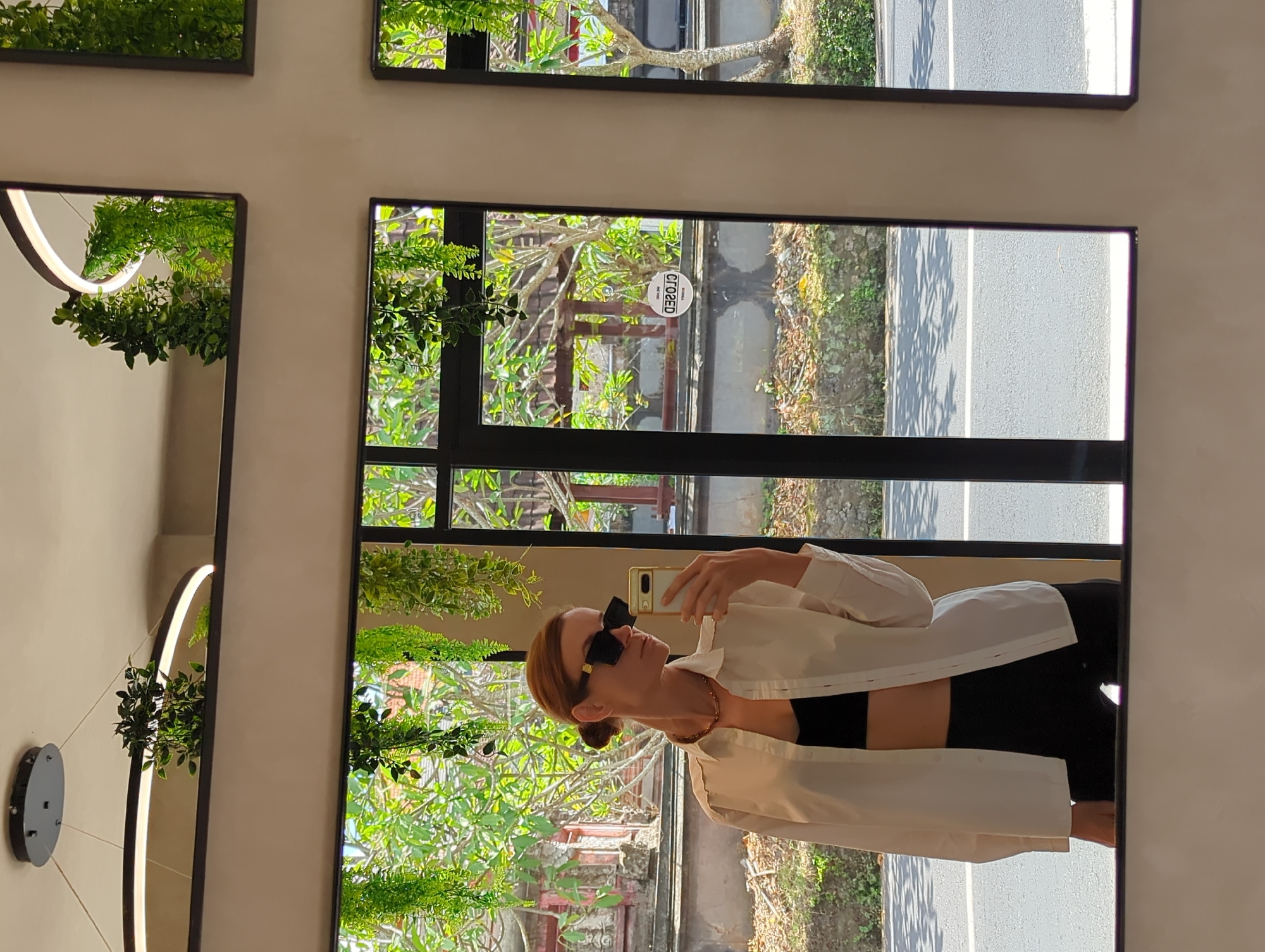 The image shows the reflection of a woman in a mirror. The woman is wearing sunglasses, a white shirt, and a black top. She is holding a phone in her hand, presumably taking a selfie. The reflection in the mirror shows a street with trees and a building. The word "CLOSED" is visible on a window. The mirrors are located in Mengwi, Indonesia.