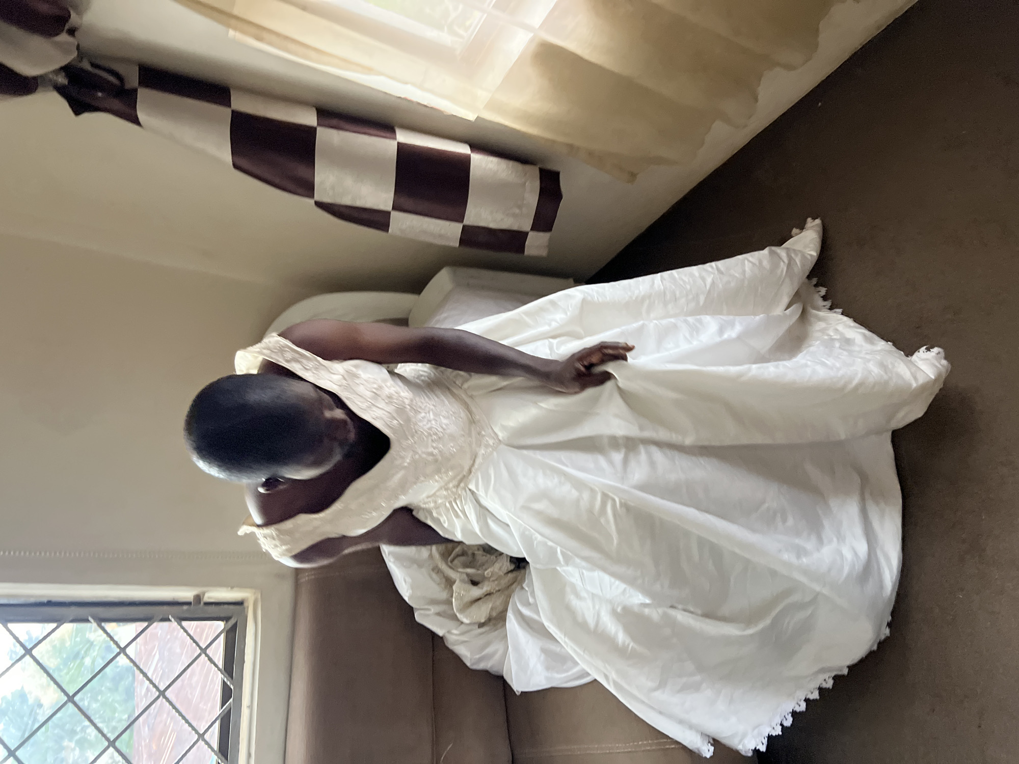 A person is sitting on a brown couch, wearing a white wedding dress. The dress has a fitted bodice with a V-neckline and intricate beading, and a full, flowing skirt with lace trim. The person is looking down, and their head is bowed. The background includes a window with a diamond-patterned grate, a wall with a checkered curtain, and another sheer curtain on the right. The lighting suggests an indoor setting, possibly a home, in Jos, Nigeria. The overall impression is one of a quiet, perhaps contemplative, moment.