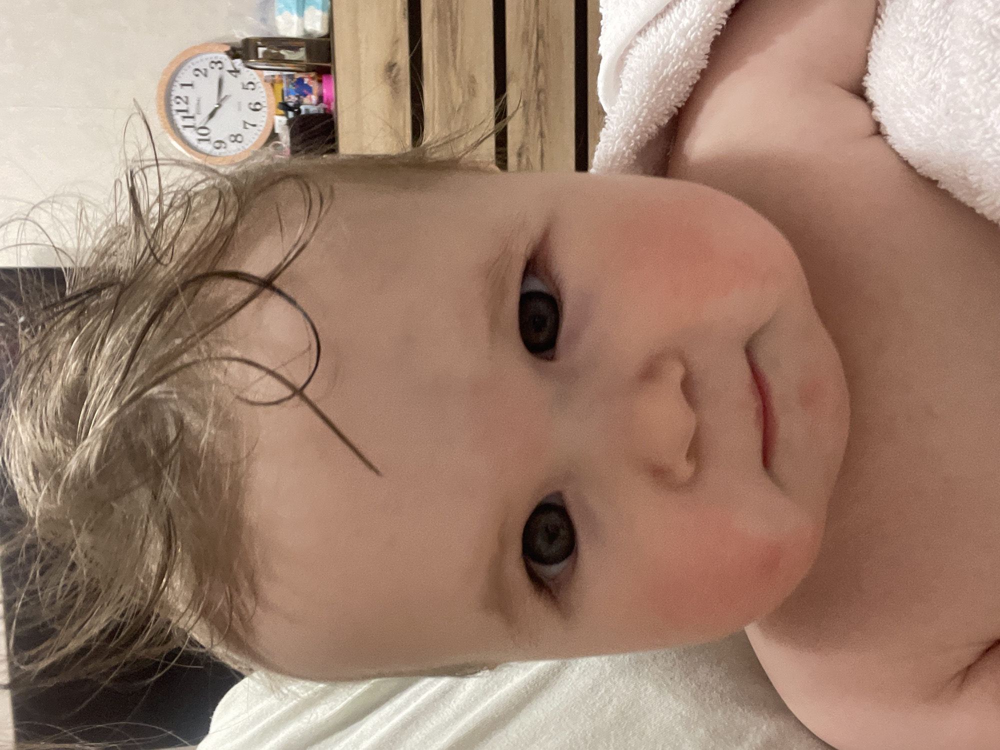 A close-up, slightly canted, indoor shot features a baby's face. The baby is fair-skinned with reddish cheeks and dark eyes. The baby's wet, blonde hair is visible at the top of the frame. A white towel is draped over the baby's body on the right side. In the background, there's a wooden-slat wall and a wall-mounted clock. The setting is not specified, and the city of Chaikovsky, Russia, is not identifiable within the visual information provided.