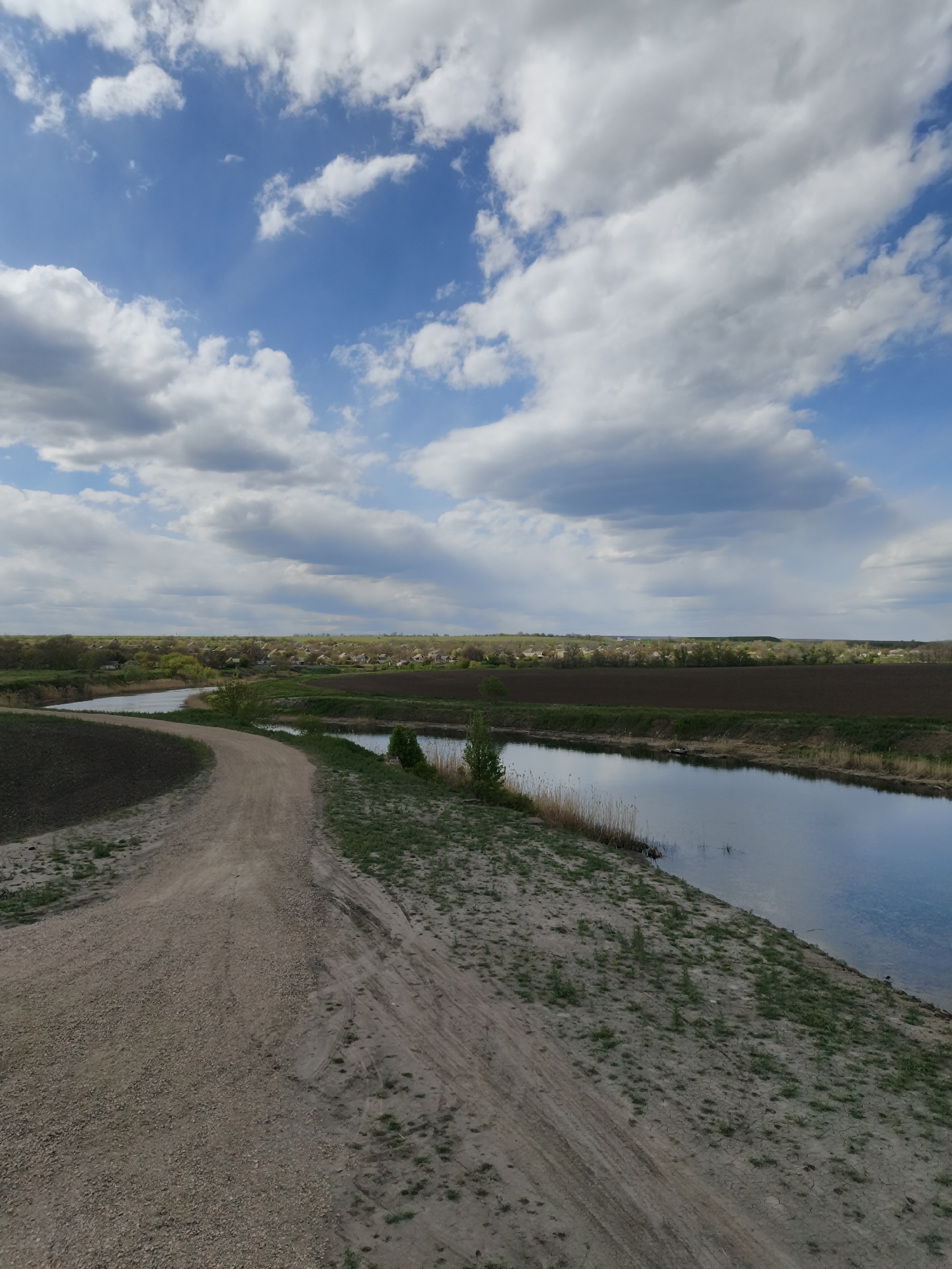 This vertical landscape image captures a serene rural scene in Inhulets, Ukraine, under a dynamic sky. A winding, light brown dirt or gravel path dominates the lower left and central foreground, with visible tire tracks suggesting recent passage. To the path's left lies a dark brown, cultivated field, while to its right, a narrow, tranquil river or canal runs parallel, its surface reflecting the sky. The riverbanks are lined with sparse green grass and reeds.
In the mid-ground, across the river and surrounding fields, a cluster of houses with light roofs indicates a distant village, partially nestled among green trees and bushes. Beyond the village, agricultural land stretches towards the horizon.
The upper portion of the image is filled with a bright blue sky adorned with an array of fluffy white and grey cumulus clouds. Patches of blue sky are visible, and subtle sun rays pierce through the cloud cover in some areas, suggesting fair weather. The overall lighting indicates it is daytime, possibly late morning or early afternoon. No people, animals, or visible text are present. The scene conveys a sense of peaceful, natural countryside.
