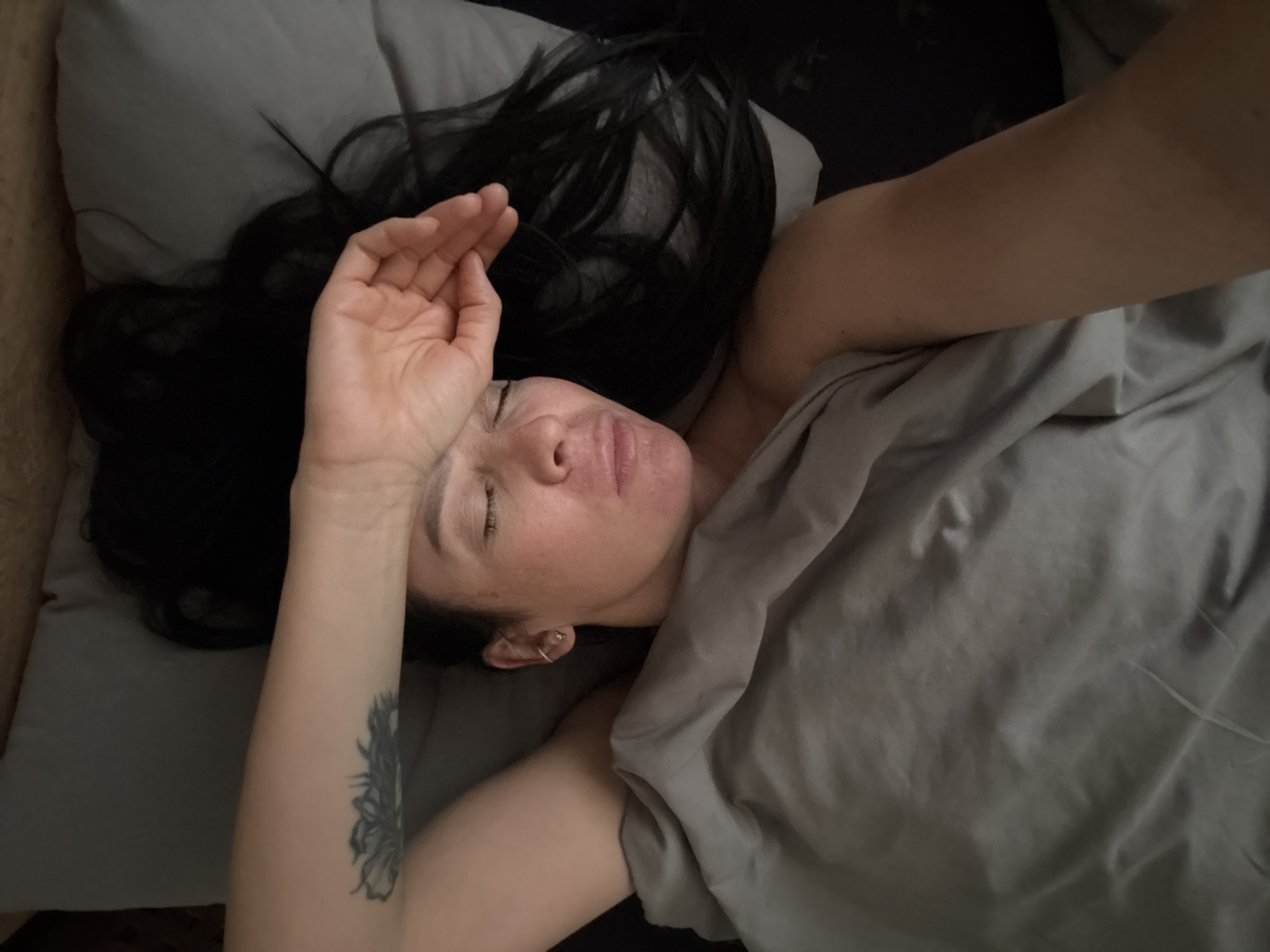 A woman with dark hair is lying in bed under a grey blanket. She has one arm extended, with her hand on her forehead. A tattoo is visible on her forearm. She is on a grey pillow. The location is unknown. The city of Chaikovsky, Russia, is not indicated in the image.