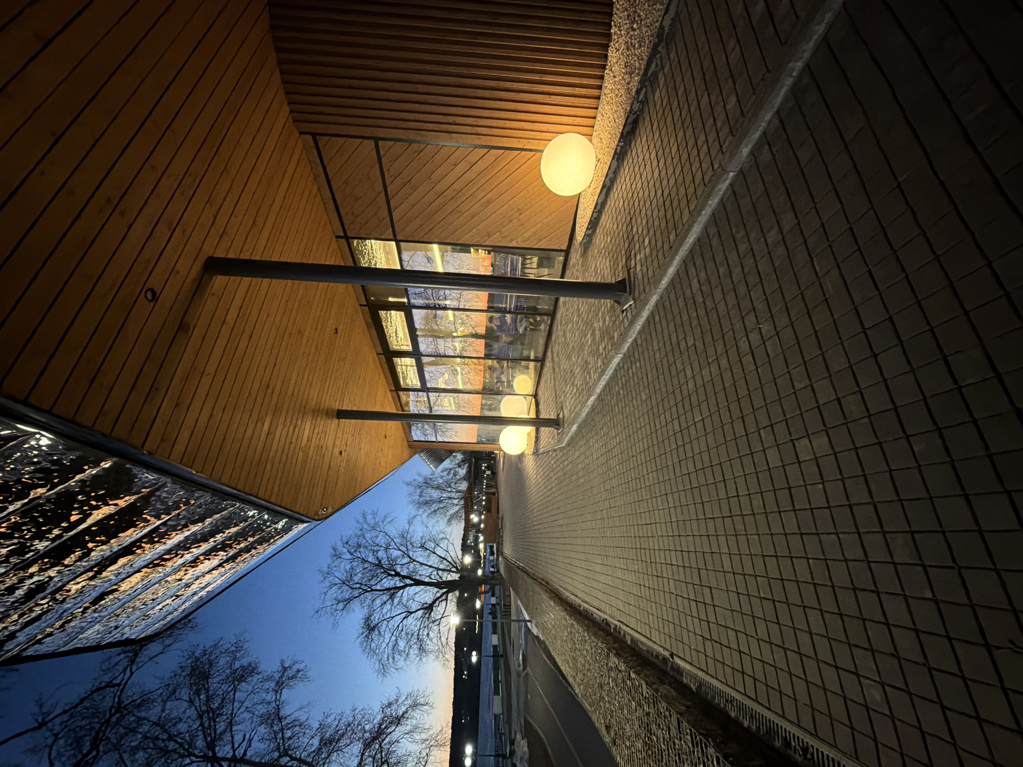The image shows an outdoor scene in Votkinsk, Russia, at dusk. The camera is tilted, creating an unusual perspective. A wooden-paneled structure with a flat roof extends along the left side of the frame. Large windows are visible within the structure, reflecting interior lights. A row of spherical streetlights illuminates a tiled walkway. A bare tree is positioned between the structure and the walkway. Beyond the tree, a street with streetlights is visible. The sky is a gradient of blue to dark.