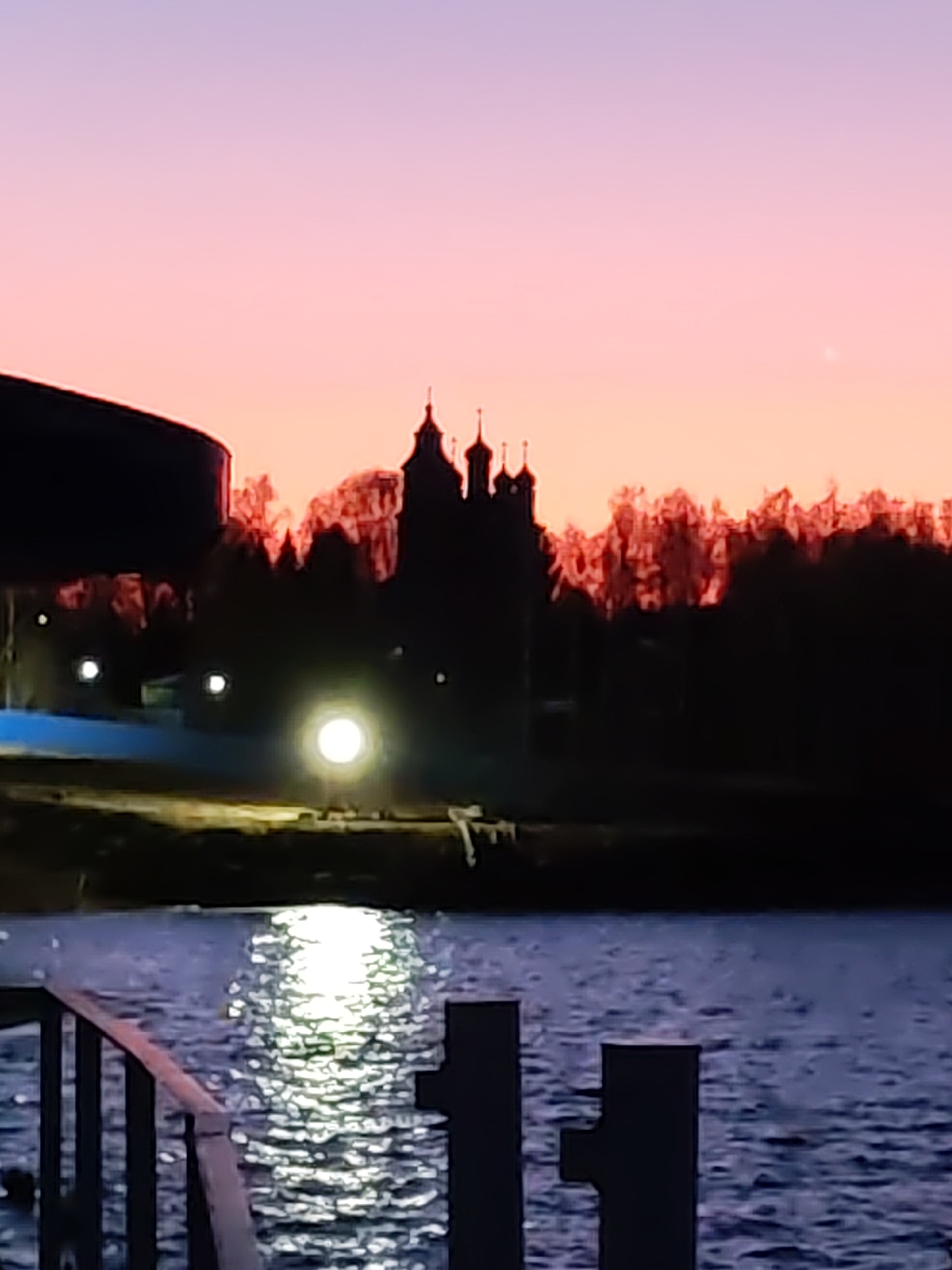 The image depicts a serene twilight landscape captured from a waterfront perspective. The sky is illuminated with a striking gradient of colors, transitioning from light purple at the top to soft pink and vibrant orange closer to the horizon, suggesting either dawn or dusk.
In the distance, a prominent silhouette of a multi-domed building, likely a church or monastery, stands against the colorful sky. Behind this structure, a dense line of trees forms another dark silhouette, with the upper edges of their foliage appearing reddish-orange, illuminated by the ambient light.
On the distant shore, a large, bright, circular artificial light source glows intensely, casting a vivid, shimmering reflection across the ruffled surface of the dark blue-grey water. Several other smaller, fainter lights are also visible along the distant shoreline. To the far left, a large, dark, curved structure, possibly part of a building or bridge, is partially visible.
In the immediate foreground, a dark metal railing or pier structure extends from the left side of the frame, while two dark, square posts rise from the water on the right. These elements suggest the photograph was taken from a dock or waterside promenade. The water itself shows small ripples and reflects the prominent light source. The scene conveys a calm and atmospheric mood. There are no people visible, and no discernible text. The weather appears clear.