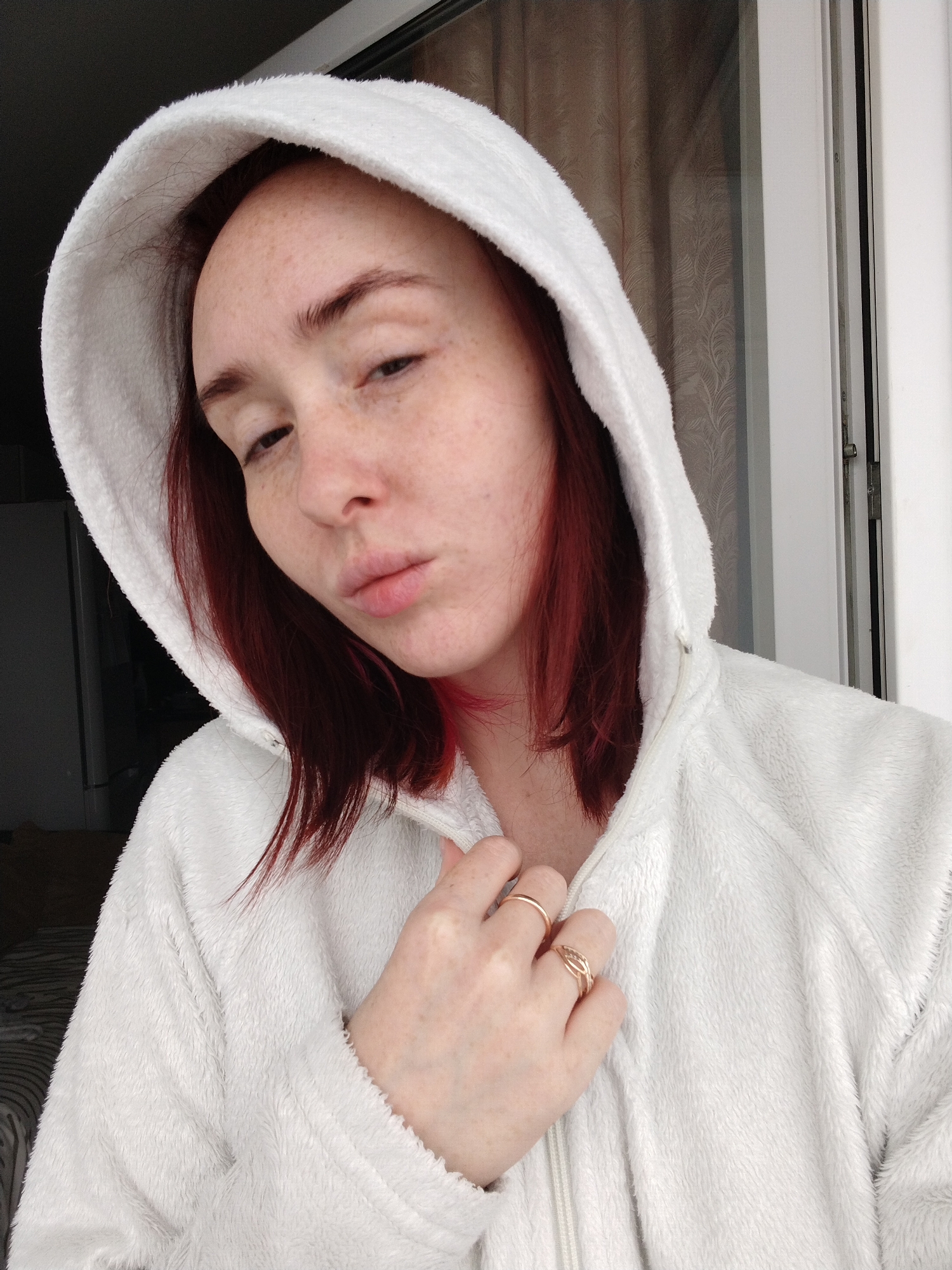 A pale-skinned woman with red hair and freckles is the primary subject of the image. She is wearing a white, fluffy hooded jacket, and her left hand is visible, adorned with two gold rings. The woman is posed indoors, presumably near a window, as indicated by the natural light and window frame visible in the background. The background also includes a curtain with a subtle pattern. The woman's expression is somewhat subdued, with a slight pout. The location in Izhevsk, Russia is not discernible from the image.