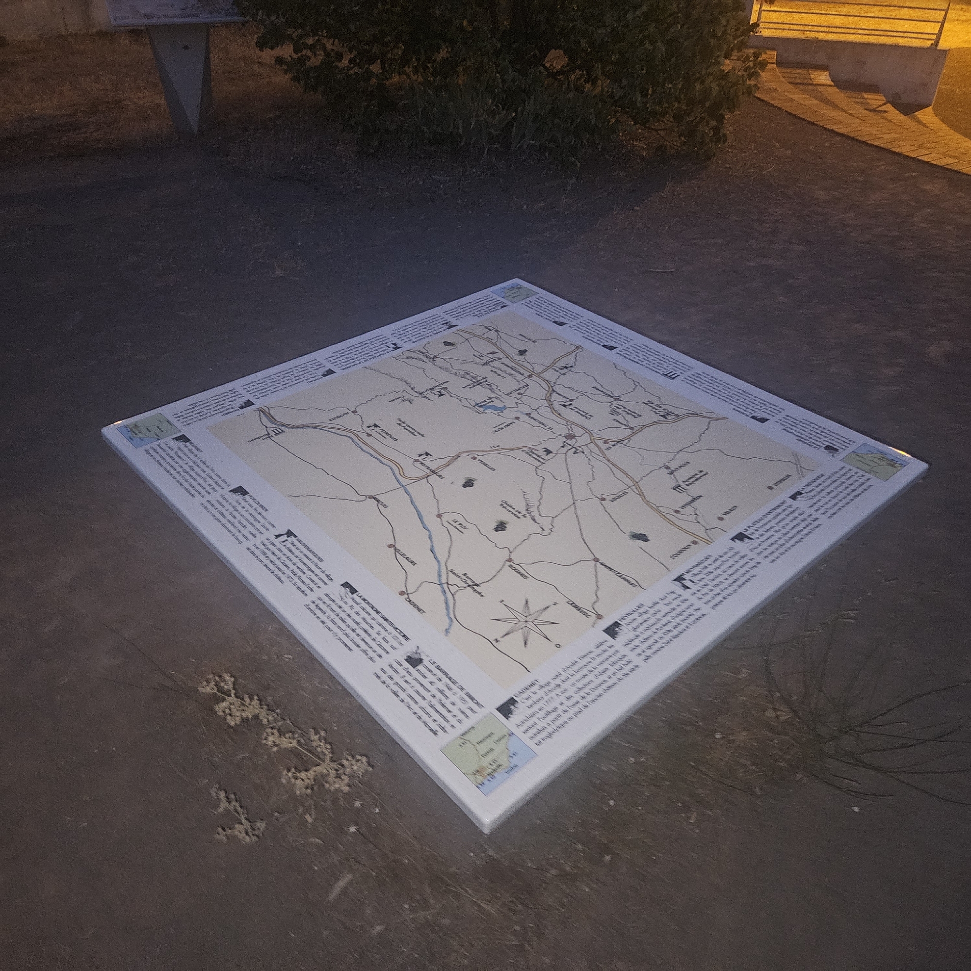 The image shows a large, square map displayed outdoors on the ground. The map is laid out flat and appears to be a tourist or informational map, depicting a region with towns, roads, rivers, and geographical features. Text in French surrounds the map, providing descriptions or historical information about various points of interest. A compass rose is visible on the map, indicating north. The ground around the map is dark and appears to be asphalt or a similar paved surface. In the background, vegetation and what looks like a brightly lit structure, possibly a building or a bench with lighting, are visible. The scene is dimly lit, suggesting it might be dusk or nighttime, with artificial light illuminating parts of the background. A small inset map is present on the lower left corner of the display. The overall impression is that of an outdoor information display in a public space, likely in Rousset, France, given the text on the map.