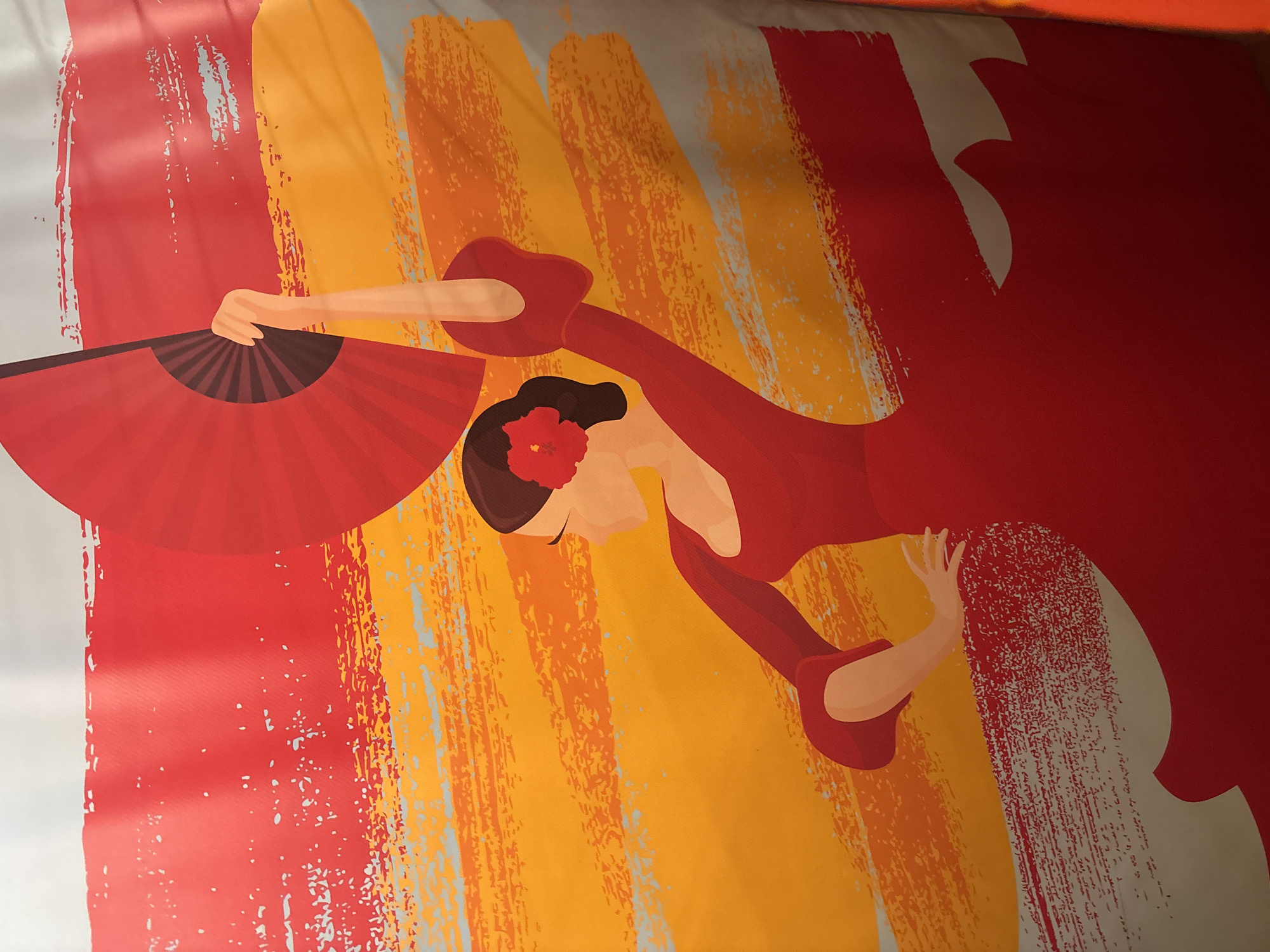 The image shows a close-up of a stylized depiction of a flamenco dancer against a background of broad, textured brushstrokes in shades of red, orange, and light gray. The dancer is wearing a red dress and holding a red fan. A red flower is in her hair. The image appears to be a section of a larger artwork or banner. There is no indication of a specific location within Dubai, United Arab Emirates.