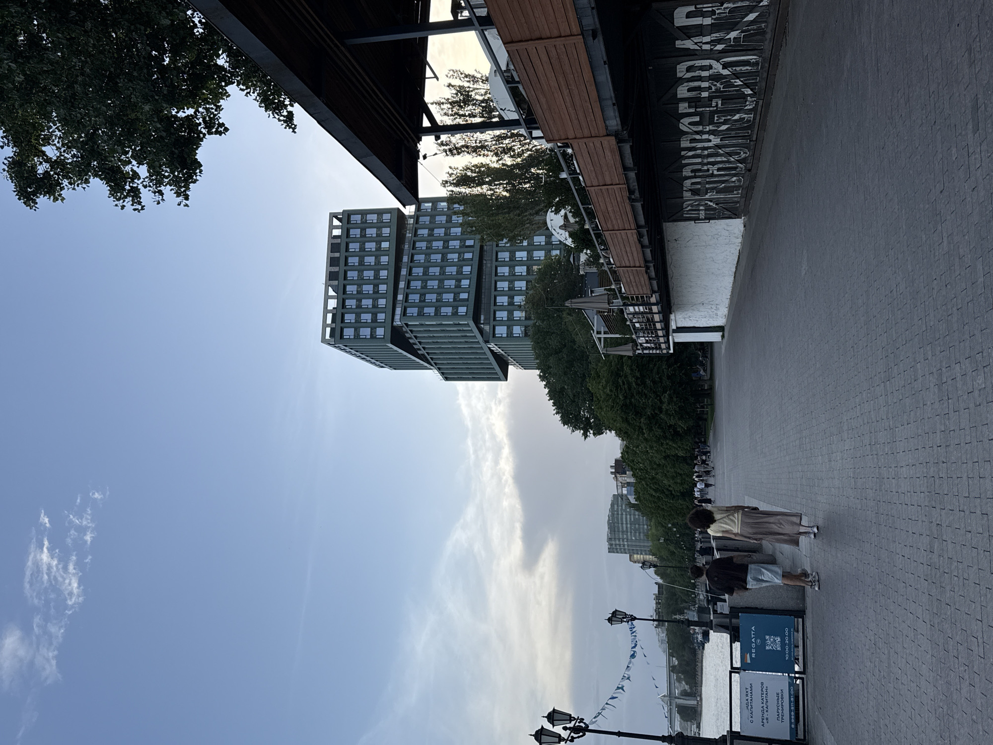 The image, taken in Rostov-on-Don, Russia, shows an outdoor scene along a paved embankment. A modern, stacked building is visible, alongside mature green trees and glimpses of the sky. The street scene features individuals strolling along the walkway, captured in the foreground are two people walking away from the camera. Streetlights and pennants are located near the river's edge. A sign advertising "Regatta" and boat rentals stands prominently on the sidewalk. A building with an awning reads "PREDBURGERRAS". The sky is overcast with wispy clouds.