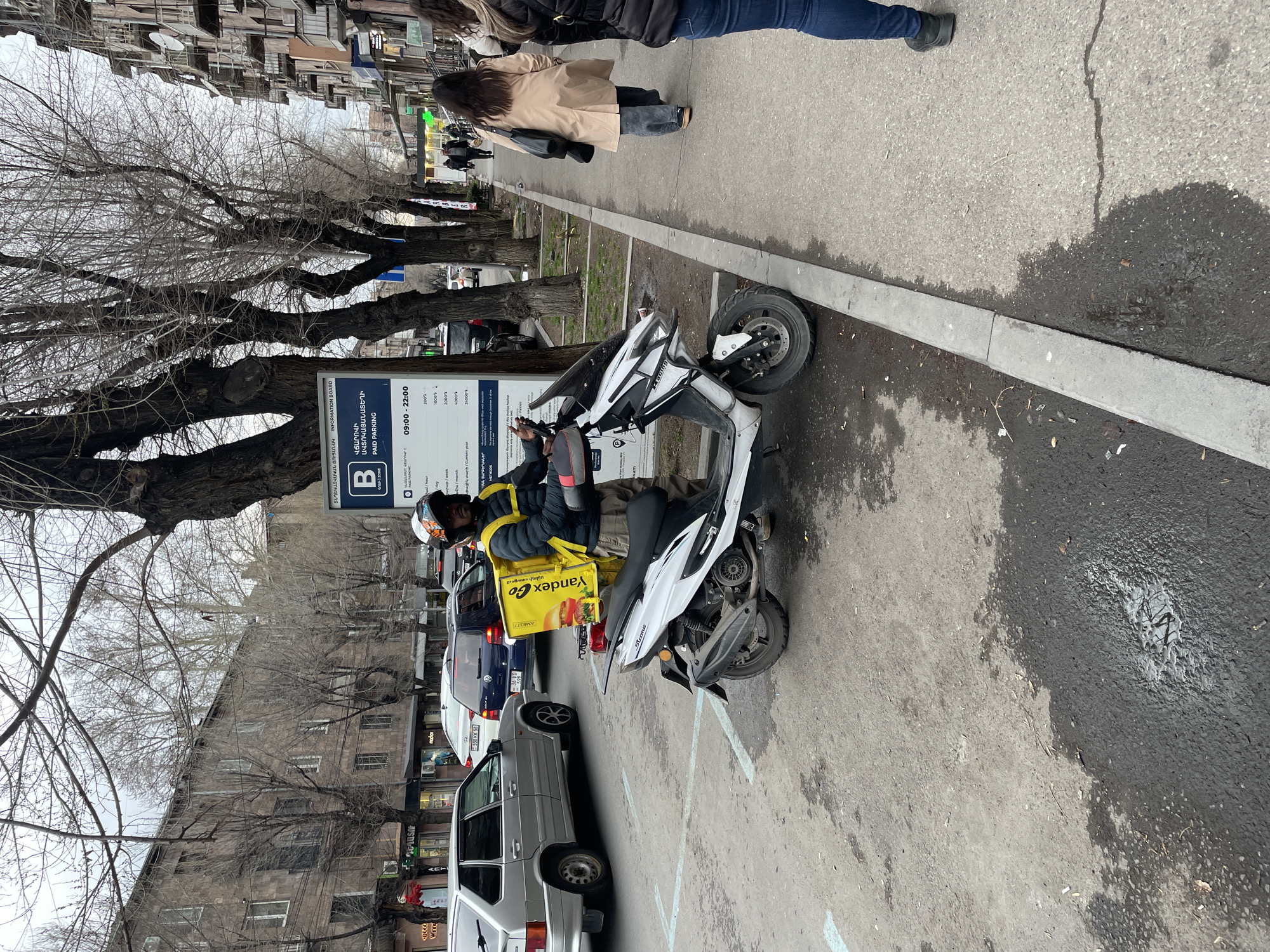 The image shows a street scene in Yerevan, Armenia, with a delivery driver on a white scooter parked in a marked parking spot on the side of the road. He is wearing a helmet and a yellow Yandex Go delivery bag. Several cars are parked behind the scooter. On the sidewalk, two people are walking. There are bare trees lining the street. A parking sign displays information in Armenian. The buildings in the background appear to be residential.