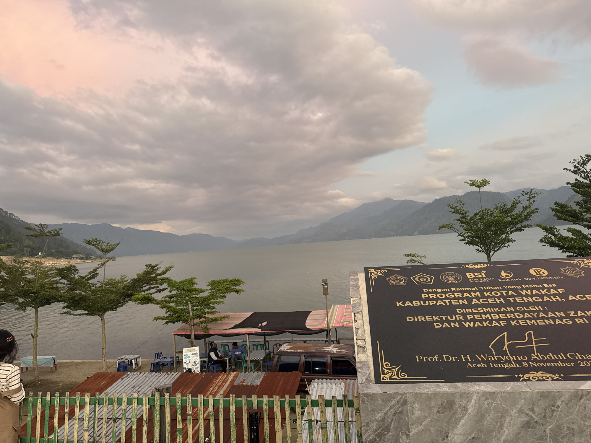 The image depicts a serene lakeside scene in Mendale, Indonesia, with a focus on a black plaque commemorating the "PROGRAM KOTA WAKAF KABUPATEN ACEH TENGAH." The plaque, adorned with gold accents and official emblems, prominently displays the inauguration date as November 8, 2020, and names Prof. Dr. H. Waryono Abdul Ghafur as the officiant.
In the foreground, a person with dark hair, wearing a striped shirt and a brown apron, stands facing away from the camera, looking towards the lake. Behind them, a rustic fence made of bamboo and corrugated metal separates the viewing area from the lakefront. Several small trees with manicured foliage are planted along the shore, adding a touch of green to the landscape.
The lake itself occupies a significant portion of the image, with calm waters reflecting the overcast sky and distant mountains. The sky is filled with dramatic, wispy clouds, some tinged with a soft pink hue, suggesting either sunrise or sunset. Several small covered stalls and tables with chairs are set up along the water's edge, indicating a recreational or dining area. A rusty brown van is parked near one of these stalls.
The background is dominated by a range of majestic, undulating mountains that recede into the distance, their peaks softened by atmospheric haze. The overall impression is one of natural beauty and a quiet, communal gathering place. The presence of the plaque suggests a significant event related to Islamic endowment (wakaf) and community development has taken place in this location.