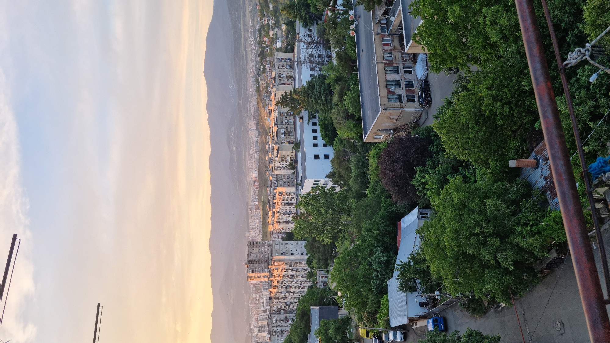 An aerial, eye-level, full shot shows a cityscape at dusk in Tbilisi, Georgia. The sky transitions from a pale blue to a warm orange as the sun sets behind distant, hazy mountains. In the foreground, a weathered, rusty metal railing runs horizontally across the bottom of the frame. Beyond the railing, the view encompasses a densely populated urban area with numerous apartment buildings and scattered trees. Some buildings are tall and imposing with balconies and numerous windows, while others are smaller, with flat roofs. Various vehicles, including cars and a blue van, are visible on the street and in parking areas. Lush green trees and foliage are interspersed throughout the scene, some obscuring parts of the buildings. The overall impression is one of a residential district bathed in the soft, golden light of sunset.