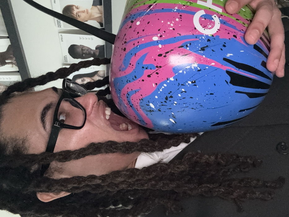 An upward-angled, candid shot captures a person with dark dreadlocks and black-rimmed glasses, positioned against a white background featuring framed images. The individual's head is tilted to their left (viewer's right), with their mouth wide open in a dramatic, possibly playful or exaggerated expression, as if yelling or laughing. A large, spherical or egg-shaped object, possibly a bowling ball, is pressed close to their open mouth. This object is vibrantly painted with a chaotic splatter design in shades of bright pink, blue, green, black, and white. A hand is visible at the top right of the image, holding the colorful object.
In the background, behind the person's head, are several framed or printed portraits on what appears to be a white wall or board. Visible text snippets next to these images include "PA" (next to a woman with short light hair), "STELLA" (below the first text, next to a person with dark skin and short hair), and "HUMAN MODES" (below "STELLA", next to another woman with dark curly hair). Part of a white letter "C" is visible on the colorful object, likely part of a brand logo. The lighting suggests an indoor setting, and the overall impression is one of a humorous or artistic interaction with the colorful object.