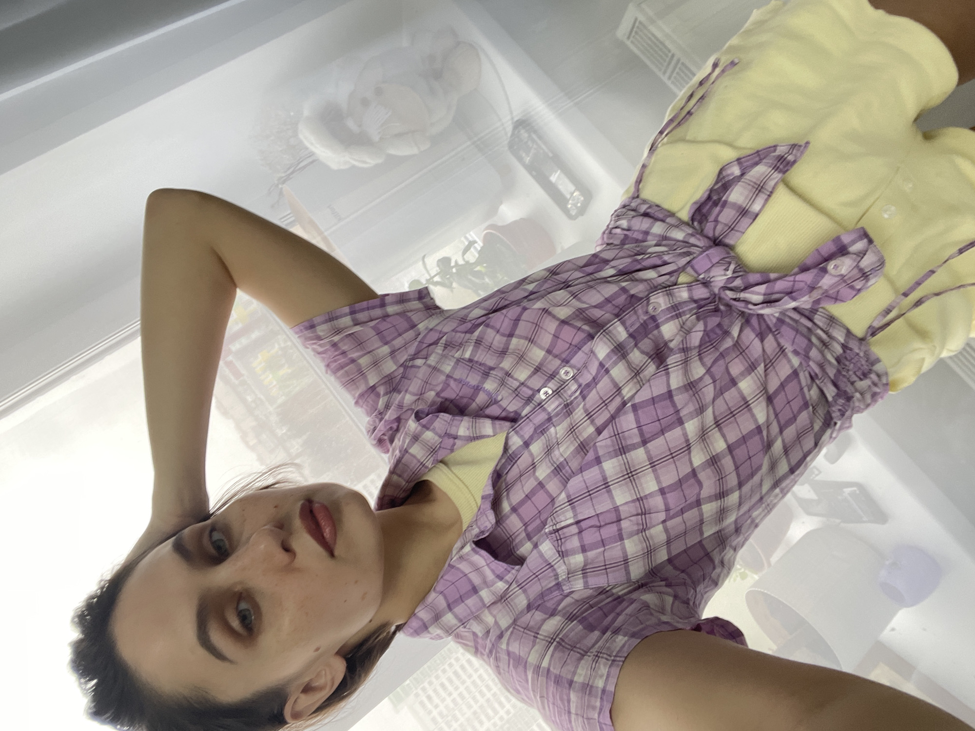 A woman is pictured in what appears to be an interior setting. She is wearing a yellow top and a purple plaid shirt tied at the waist. The background contains indistinct objects that resemble a teddy bear, plant, and various containers, which are likely household items. Based on the visible details, it is not possible to determine the location in the city of Izhevsk, Russia.
