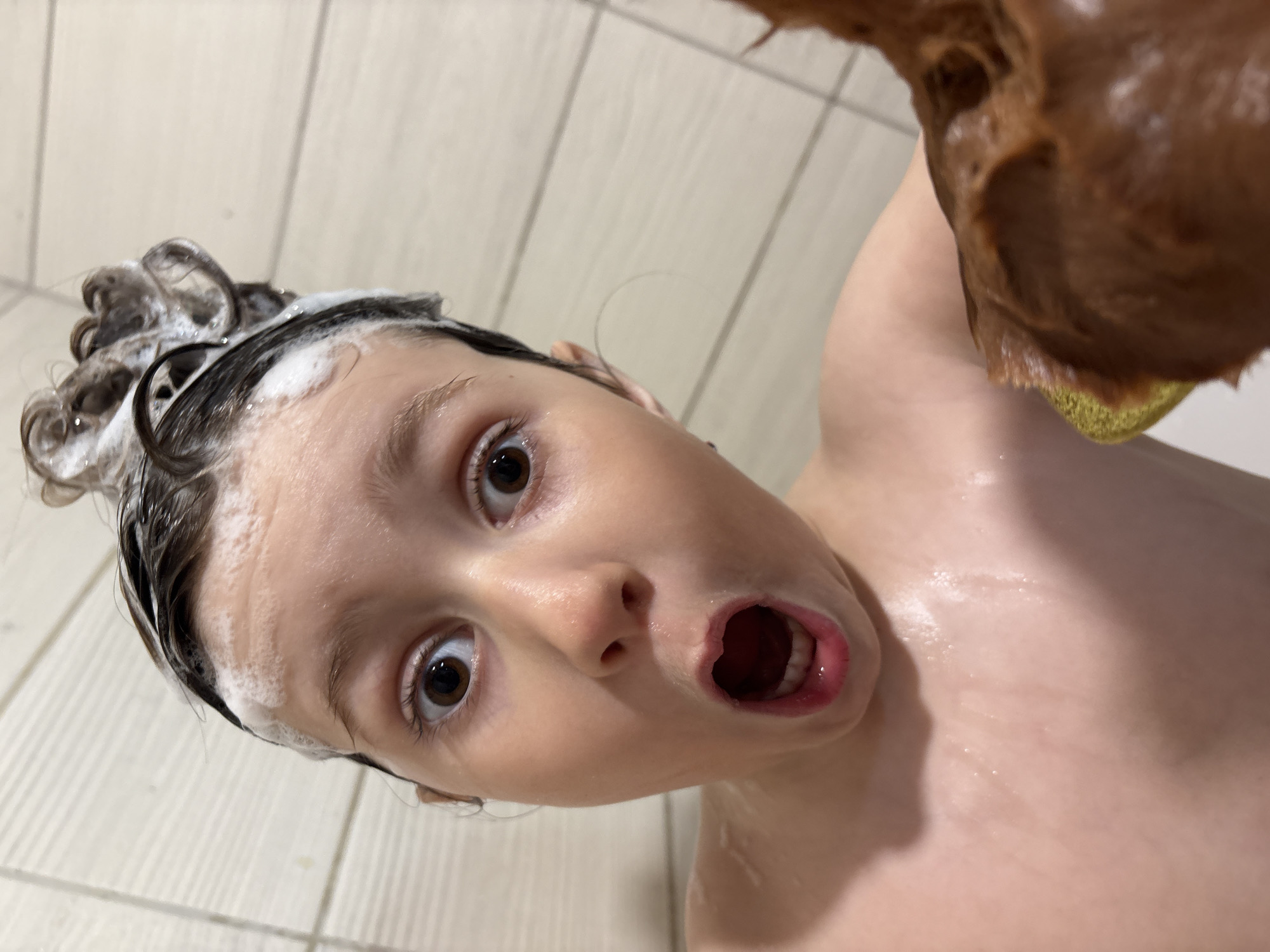 A young individual with soap in their hair is in a shower with their mouth open. The person is holding a brown, porous object. The location is a shower with white-tiled walls. The location in the city of Chaikovsky, Russia, cannot be determined from the image.