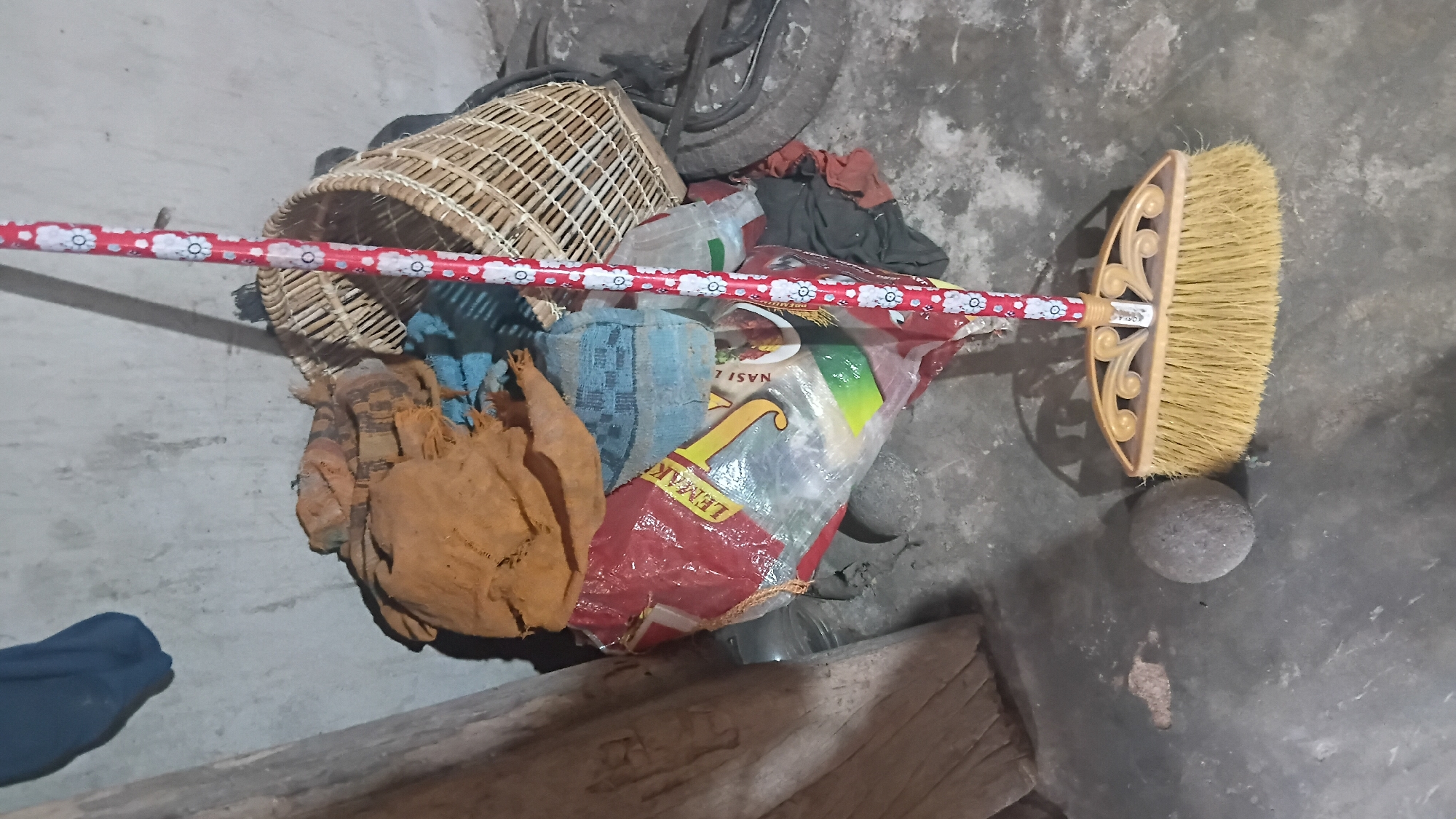 A broom with a red and white floral pattern on its handle is positioned in the foreground, with its bristles resting on a concrete floor. To the left of the broom is a round, gray stone. Behind the broom and stone, a pile of items is visible, including a woven basket, various fabric scraps in brown and blue hues, and a large plastic bag with red and yellow labels that appear to be in Indonesian. A portion of a bicycle wheel is visible on the right side of the image. The background consists of a textured, light-colored wall. The overall impression is that of a storage area, possibly a corner of a home or an outdoor workspace. The scene is set in a rustic environment, with the concrete floor and textured wall suggesting a non-urban or semi-urban setting. It is not possible to determine the specific location within the city of Prabumulih, Indonesia from the image alone.