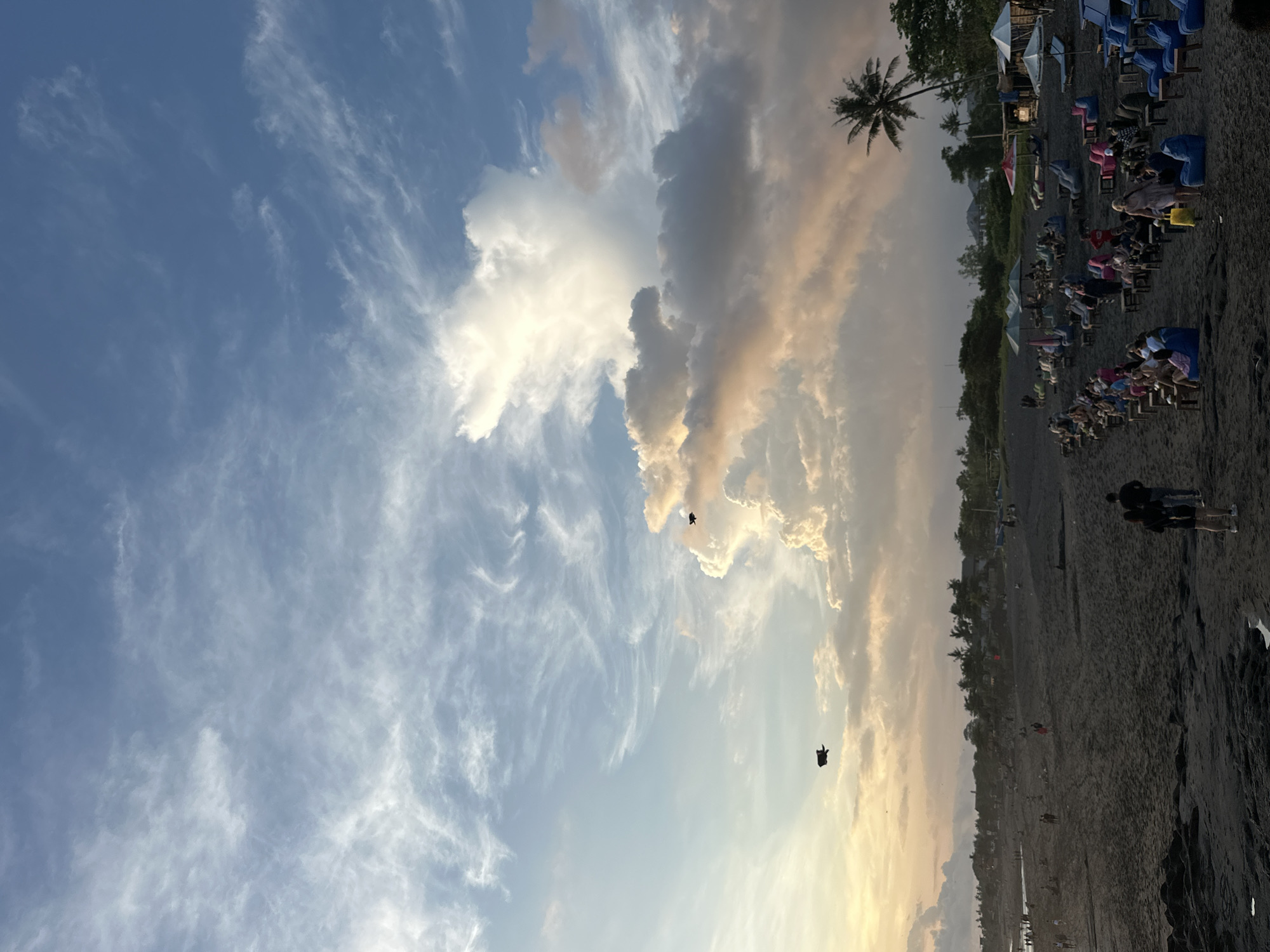The image shows a beach scene under a partly cloudy sky, oriented in a vertical format. The beach appears to have dark sand. Along the edge of the beach are rows of chairs and umbrellas. People are visible sitting in the chairs. Two figures are standing near the edge of the frame. Overhead, two objects, possibly kites or similar, are suspended in the sky. A palm tree is visible amidst the seating area. The location is Unknown, Unknown.