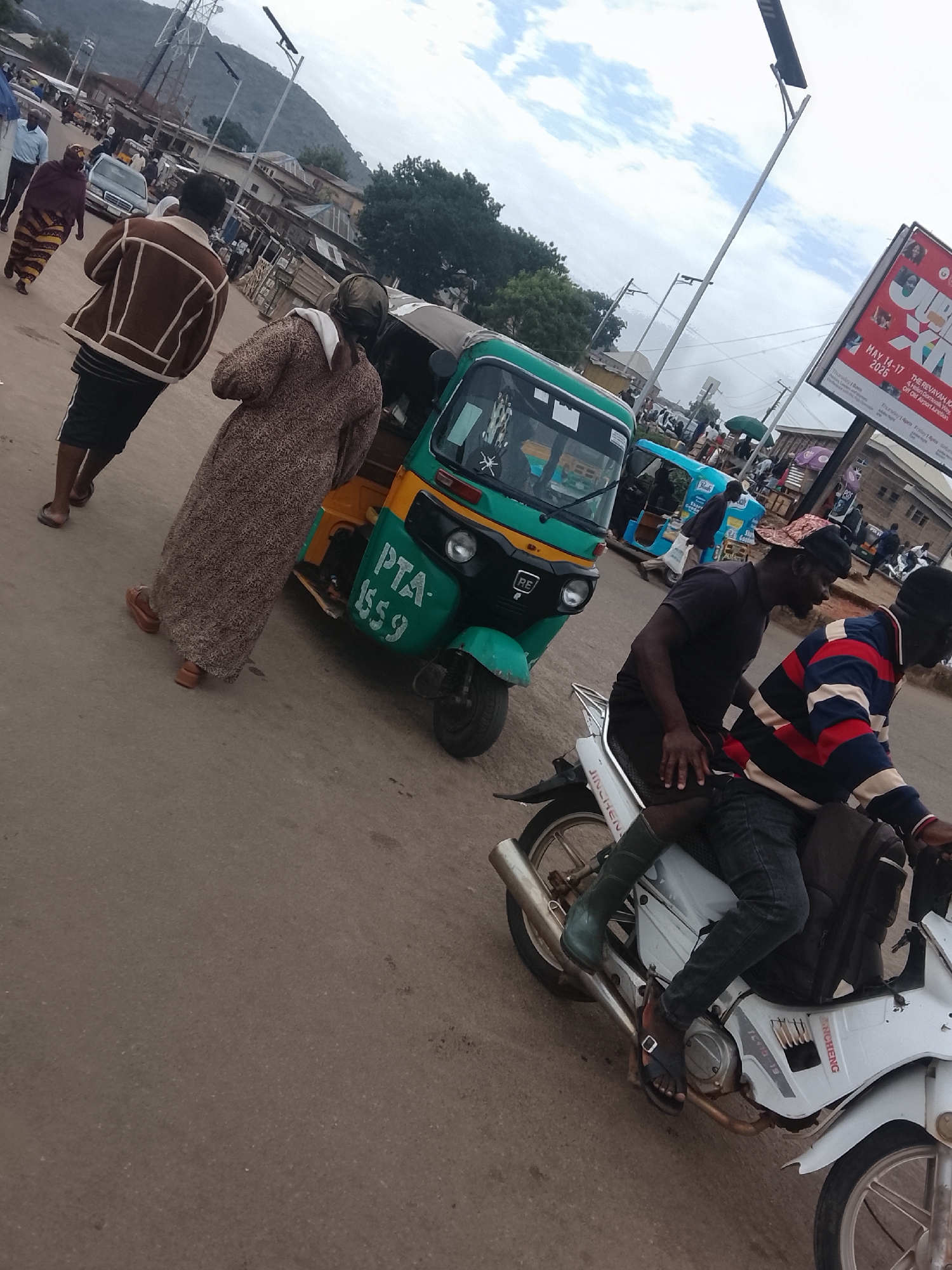The image captures a street scene in Gwong, Nigeria. An auto rickshaw, primarily green and yellow, is positioned near pedestrians. One individual wears a brown-and-white jacket and shorts, and another wears a long, patterned dress. A motorcycle, white in color, carries two men. One man wears a black shirt and the other wears a striped sweater. A large advertisement sign is visible in the background, indicating an event from May 14-17, 2026. Other individuals, cars, buildings, and power lines are visible in the background, along with a hill in the distance. The sky is overcast.