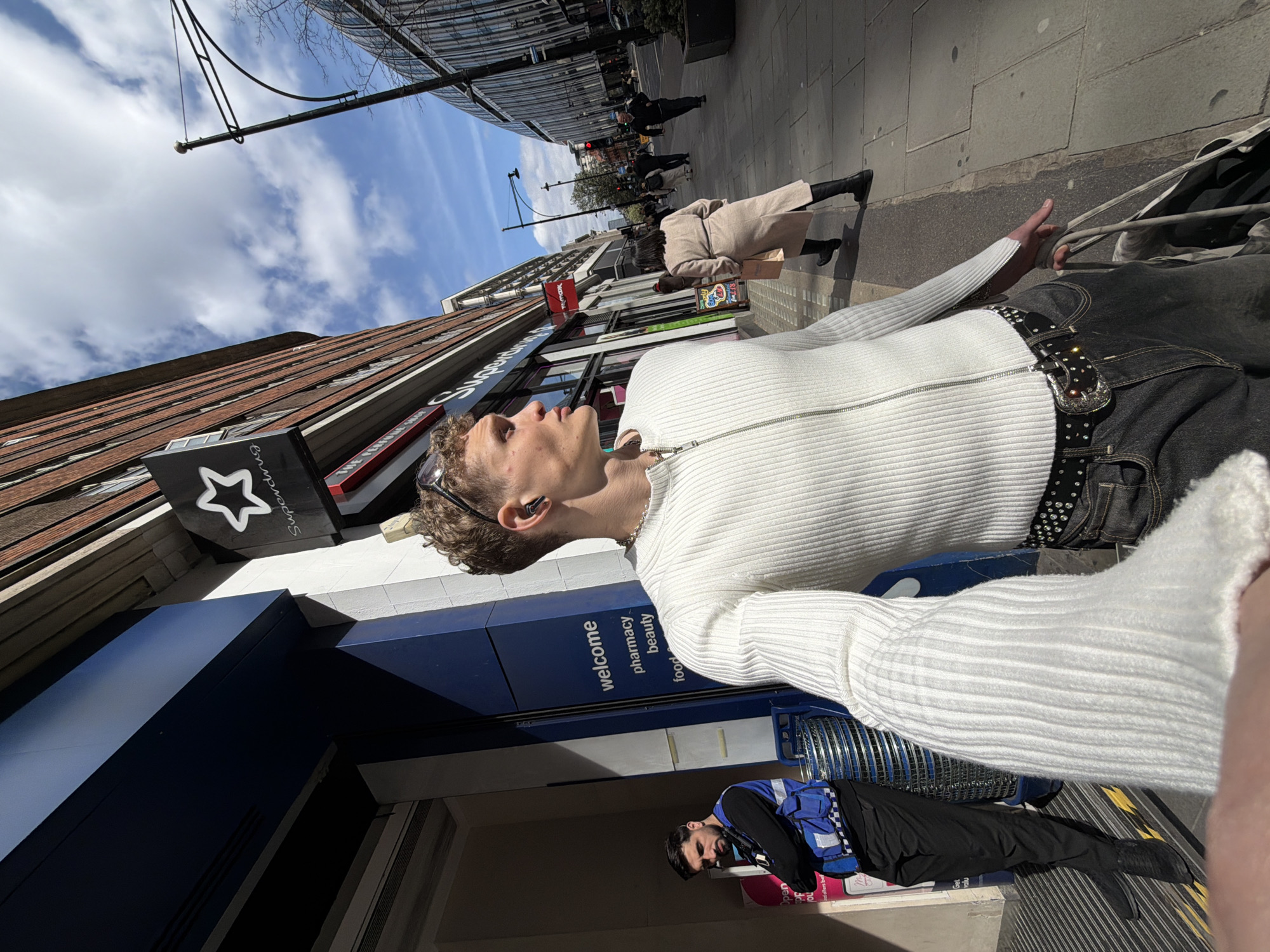 The image depicts a street scene in London, United Kingdom. A young adult male, fair-skinned with curly brown hair and wearing sunglasses on his head and an earbud in his ear, is the foreground subject. He is wearing a white, ribbed long-sleeved shirt with a zipper and a studded belt over dark-colored jeans. He is standing next to a shopping cart.
Behind him is a storefront with a blue and white sign that reads "welcome pharmacy food beauty". A man in a black jacket and blue vest lies on the ground next to a shopping cart. Pedestrians walk along the sidewalk in the background, with buildings lining both sides of the street. The sky is partly cloudy. The street appears to be a typical urban shopping area.