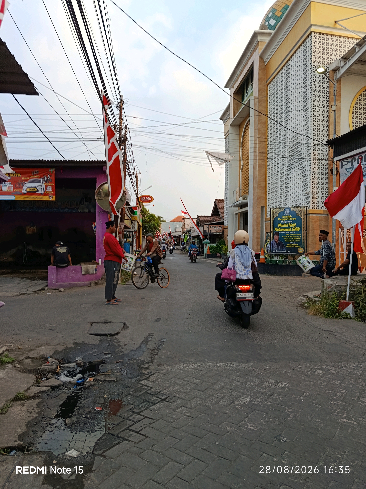 The image depicts a street scene in Keboansikep, Indonesia, possibly during a celebration or cultural event, as indicated by the presence of Indonesian flags (red and white) decorating the area. The street is paved with interlocking blocks. On the right side of the street is a building with yellow and white facade, featuring an arched window and decorative perforated walls, suggesting it might be a mosque or a similar public building. A banner with a portrait and text in Indonesian is displayed on this building.
On the left side of the street, a purple building with a metal roof is visible, with advertising banners hanging on its facade. A man in a red long-sleeved shirt and grey pants is standing near a bicycle, talking to another person. Several motorcycles are present on the street; one is being ridden by a person wearing a white helmet and a light blue jacket. Another motorcycle is parked in the foreground.
There are power lines crisscrossing overhead, a common feature in urban and semi-urban environments in Indonesia. On the ground, near the purple building, there is a drain or pothole filled with murky water and debris, including plastic bags and other refuse. A man is seated on a low wall in front of the purple building. Another man is seated on a concrete structure on the right side of the street, holding a white bucket.
The sky is overcast, with the time stamp "28/08/2026 16:35" visible in the bottom right corner, and "REDMI Note 15" in the bottom left, indicating the image was taken with a Redmi Note 15 phone on August 28, 2026, at 4:35 PM.