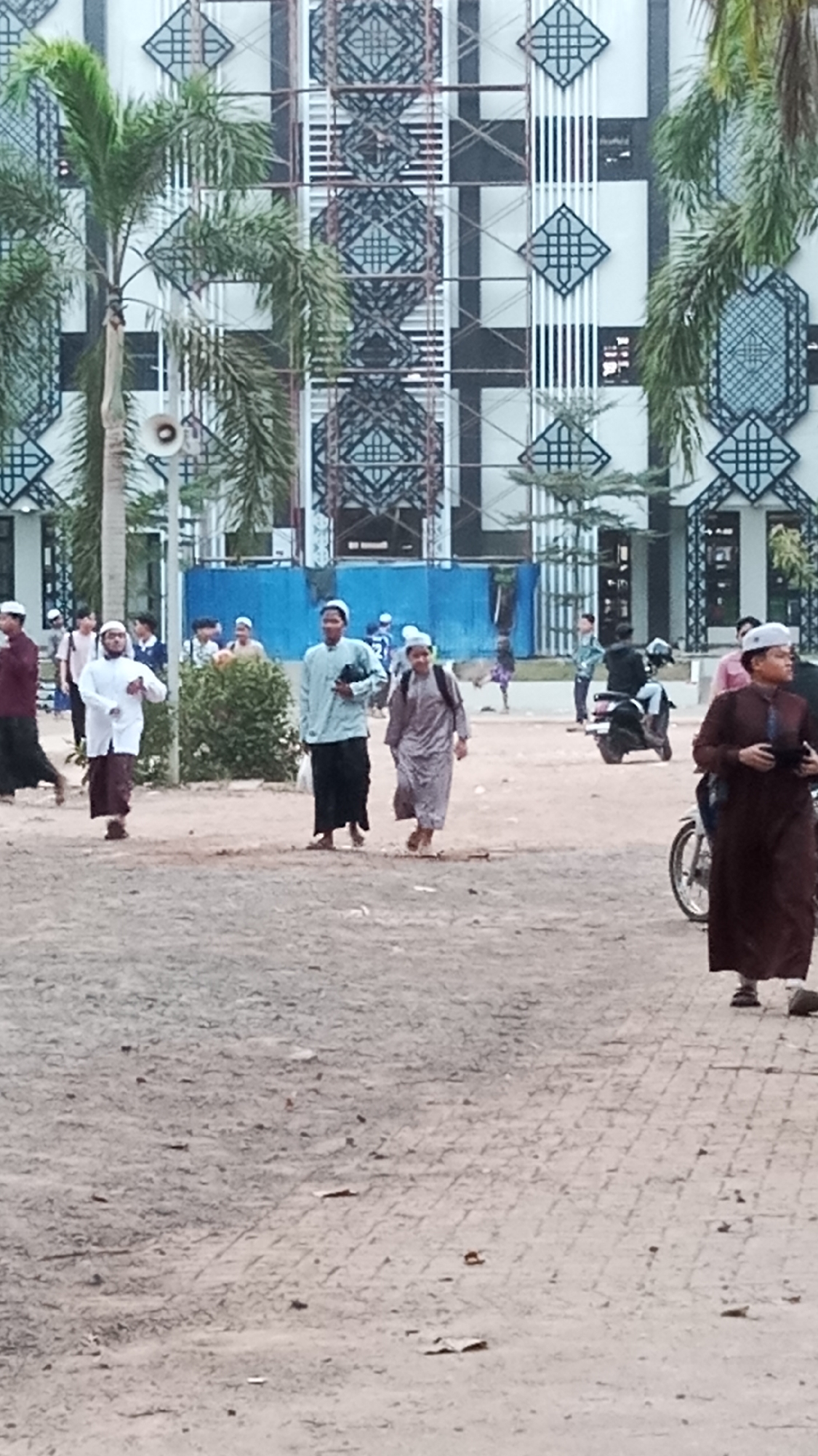 A group of young men, most wearing traditional Islamic attire consisting of robes and skullcaps, are walking on a dirt and paved pathway in front of a modern building with intricate geometric patterns on its facade and palm trees on the left. The building appears to be a school or religious institution. In the background, a blue tarp is visible, suggesting ongoing construction or a temporary structure. A motorcycle is parked to the right, with one person seated on it. The overall scene depicts a casual gathering or movement of people outdoors. The location is identified as Tanjung Rema, Indonesia.