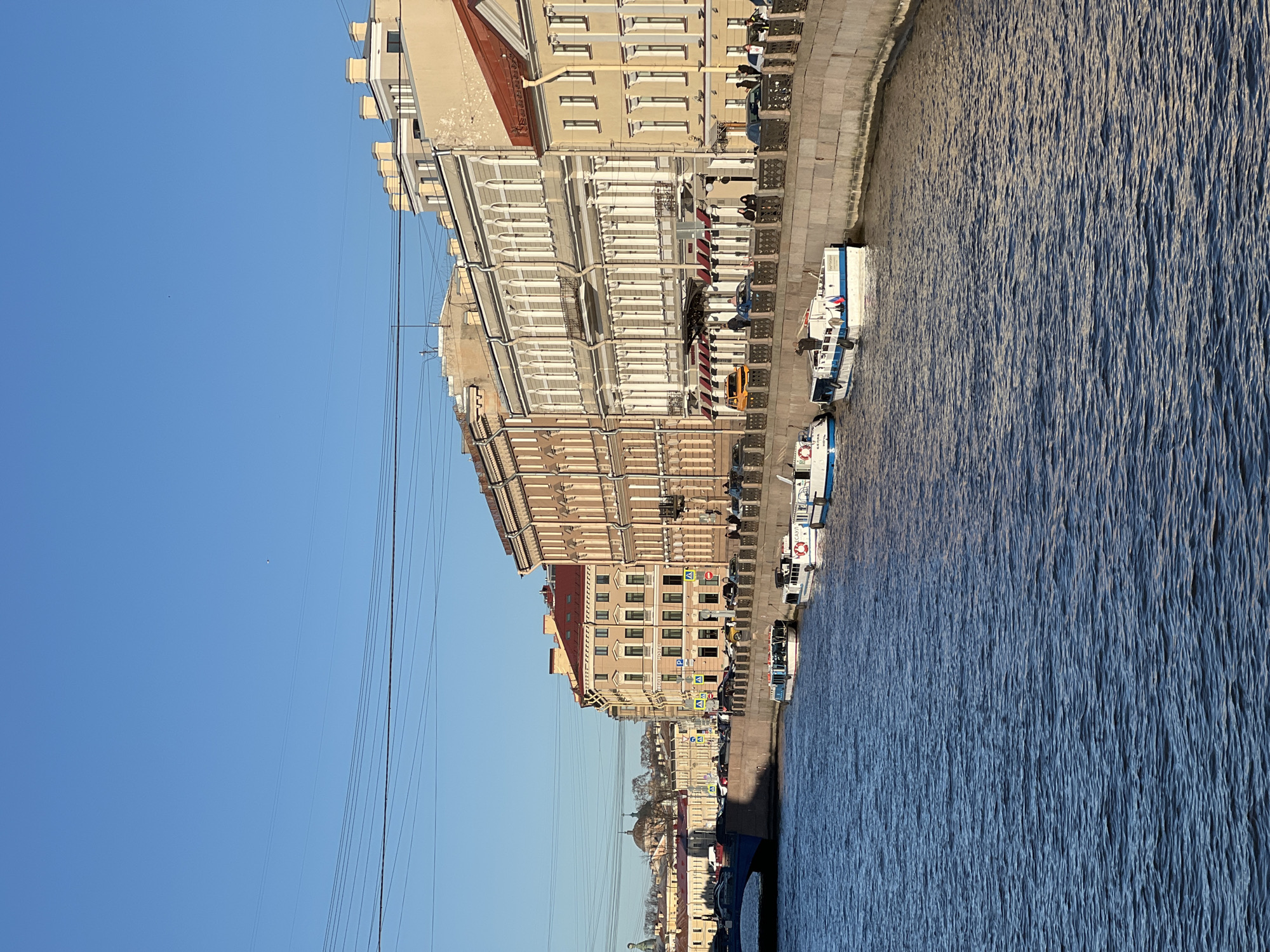 This image captures a view of a section of the Fontanka River embankment in Saint Petersburg, Russia. The river flows to the right, its surface marked by distinct ripples. Along the left bank, a row of buildings lines the embankment, exhibiting a variety of architectural styles and colors, from light beige to deeper shades of terracotta. Several boats are docked along the embankment, adjacent to a paved walkway where parked cars and sporadic pedestrian activity are visible. Overhead power lines stretch across the upper left, creating a network against a clear blue sky. Further down the embankment, the skyline of Saint Petersburg emerges with identifiable landmarks, including domes and spires. The overall atmosphere is sunny and calm.