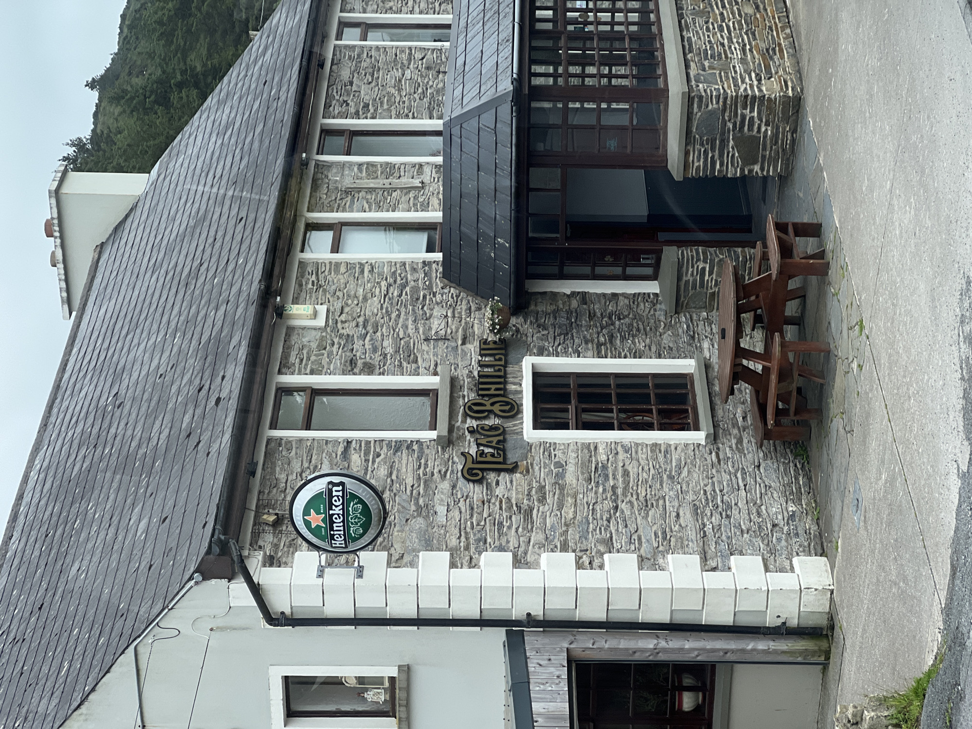 The image depicts the exterior of a stone building in Gortahork, Ireland. The building appears to be a pub or restaurant, with a wooden sign displaying "TAEG SHILL" in gold lettering. A prominent Heineken beer advertisement is also visible. The architecture features a grey slate roof and rough-hewn stone walls. White decorative crenellations run along the lower section of the building. A wooden picnic table and benches are set up outside, suggesting an outdoor seating area. The scene is set against a backdrop of lush green trees and an overcast sky, hinting at a cool, possibly damp day. The image is taken from a slightly elevated perspective, looking towards the building. There are no people visible in the frame. The overall impression is one of a traditional establishment in a rural Irish setting.