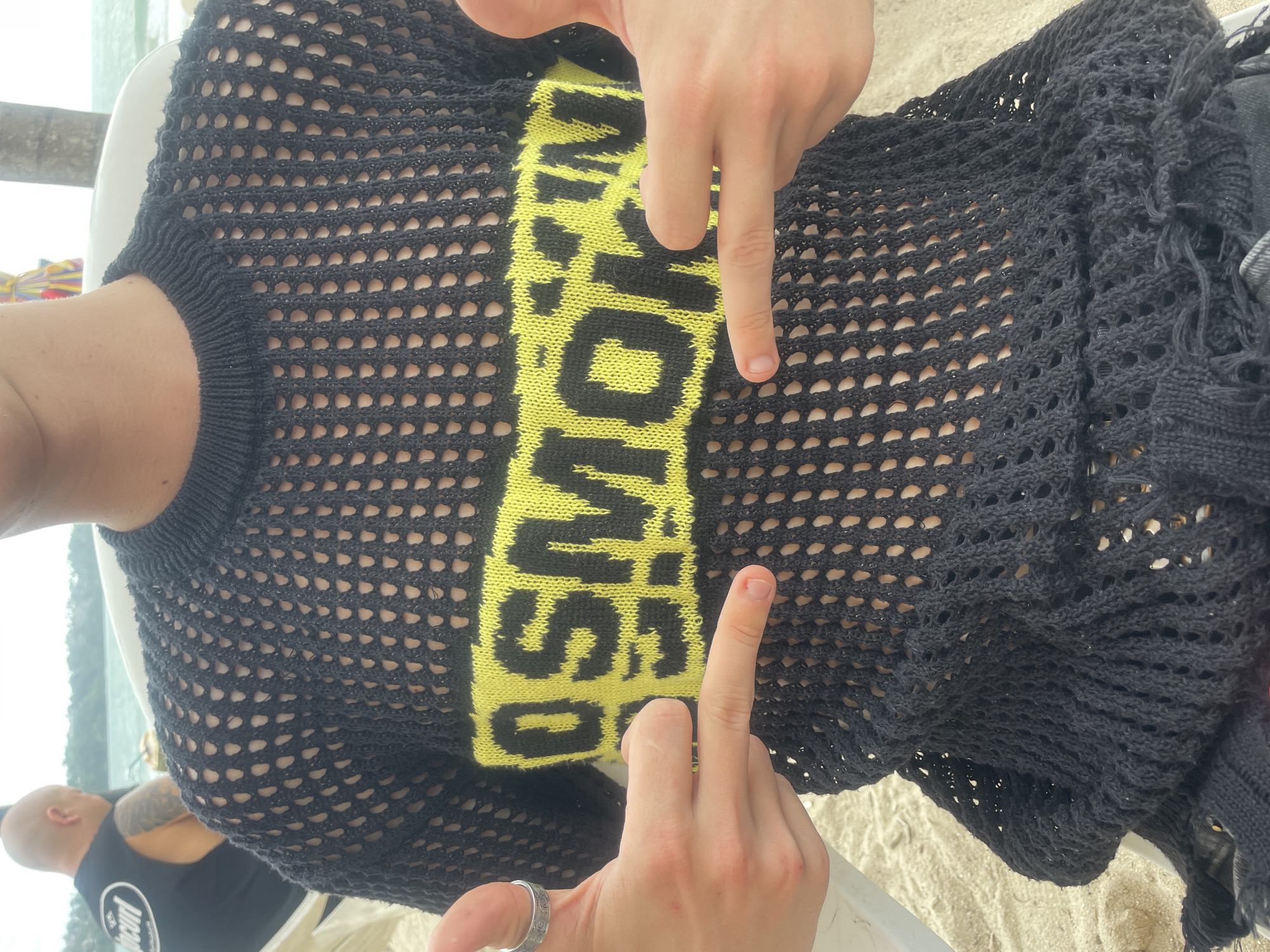 The image depicts a person wearing a black, open-weave, knitted top with a yellow and black band across the chest, featuring the word "SMOKING" in large letters. The person is pointing at the top with both hands. A ring is visible on the left hand. In the background, sand and a partially visible person with tattoos are present, suggesting a beach setting. The location is Choeng Thale, Thailand.