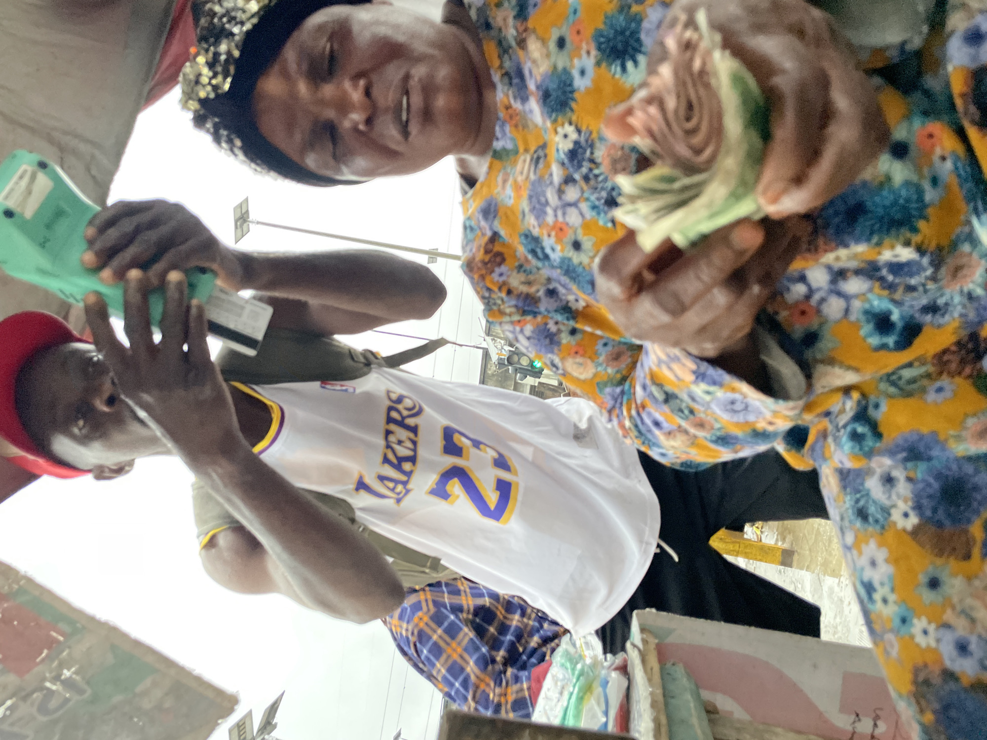 In Lagos, Nigeria, a young man wearing a red baseball cap and a white Lakers jersey with the number 23 is shown holding a green electronic device, possibly a point-of-sale terminal, in his right hand. He is positioned behind a woman who is counting a wad of cash in her hands. The woman is wearing a colorful floral print dress and a black headscarf with sequined embellishments. The scene appears to be outdoors, possibly at a market or street vendor stall, with a blurred background suggesting an urban environment. A second person in a blue and orange plaid shirt is partially visible to the left. The overall impression is a transaction taking place in a busy, everyday setting within the city.